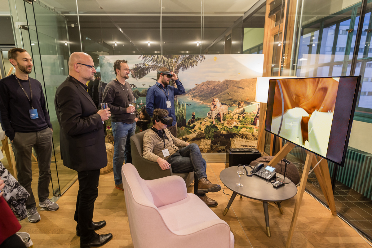 Vier Personen stehen in einem Raum und beobachten einen Bildschirm, wo sie sehen, was eine fünfte Person mit VR Brille sieht.