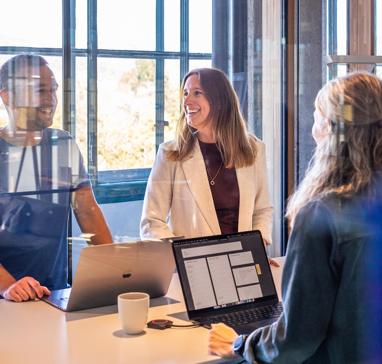 Drei Kollegen unterhalten sich freundlich an einem Stehtisch mit Laptops in einer lichtdurchfluteten Arbeitsumgebung.