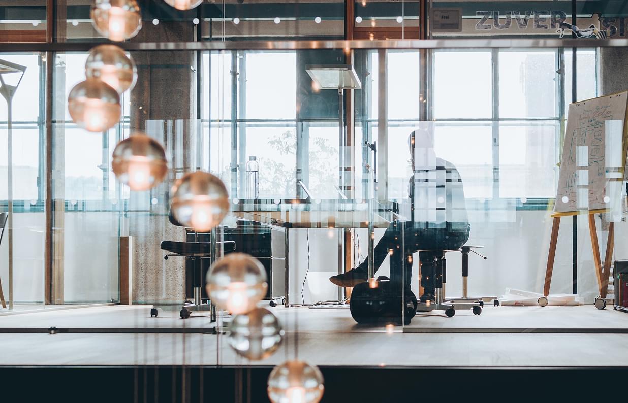 Blick durch eine Glaswand mit dekorativen Leuchten auf eine Person, die konzentriert an einem Schreibtisch arbeitet