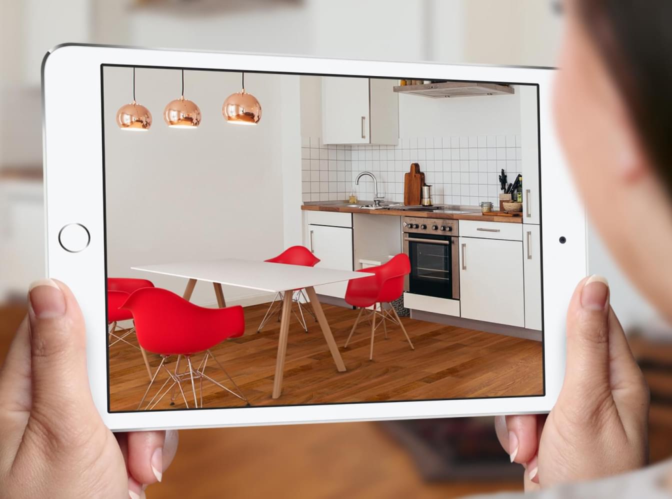 Tablet zeigt mit AR eingerichtete Küche mit weißen Möbeln, roten Stühlen und Kupferlampen über Holzboden.