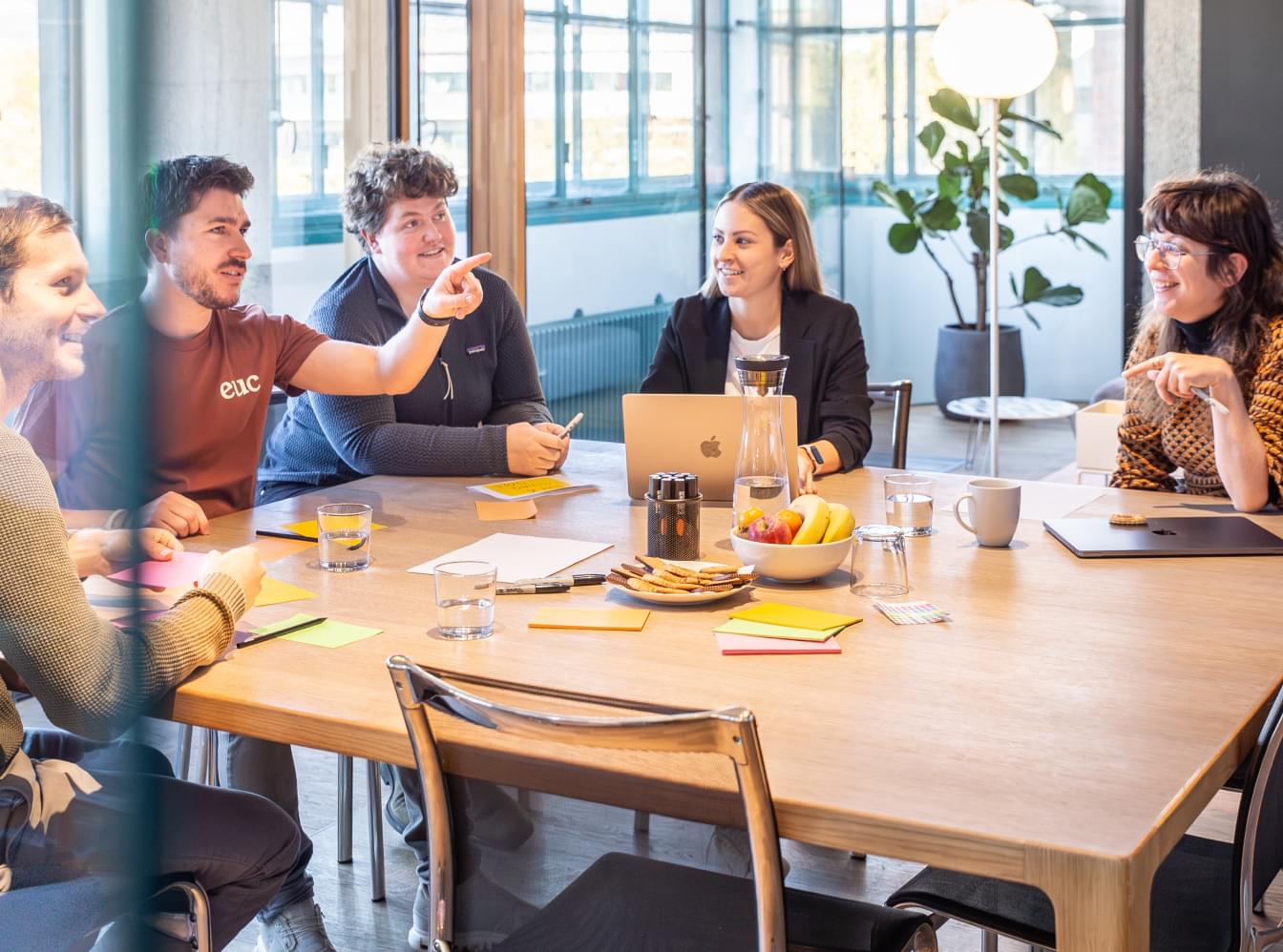 Ein Team bespricht Pläne an einem Holztisch mit Laptops und Obstkorb. Der Blick geht durch eine Glaswand, die das helle Büro reflektiert.