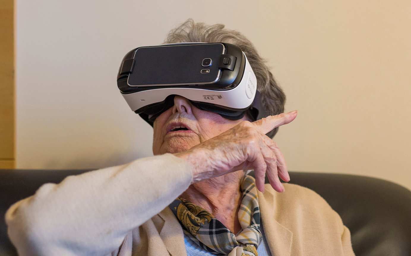 Ältere Frau mit VR-Brille sitzt auf dem Sofa und zeigt beeindruckt in die Luft.