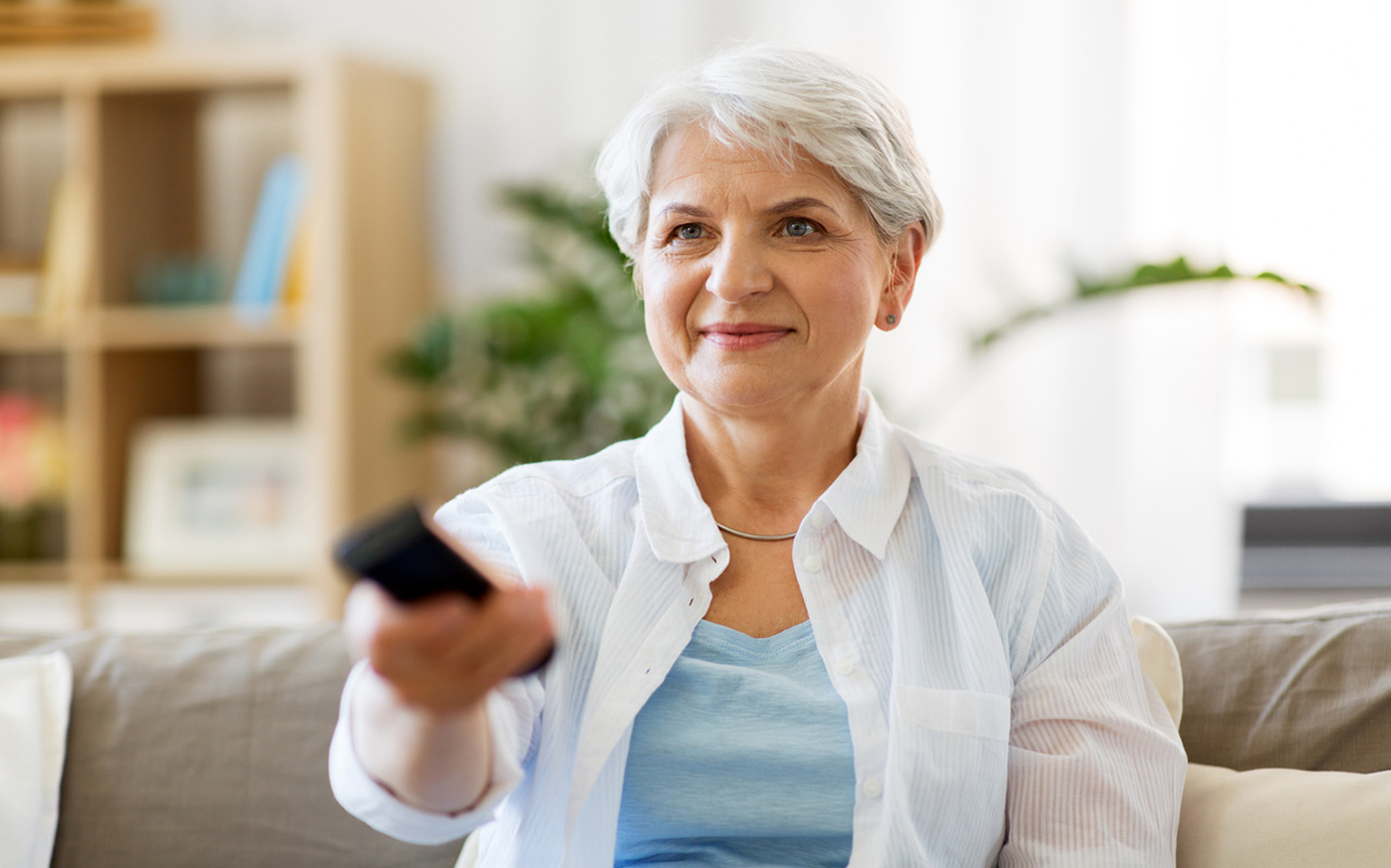 Ältere Frau sitzt auf dem Sofa und lächelt, während sie mit einer Fernbedienung den Fernseher einschaltet.