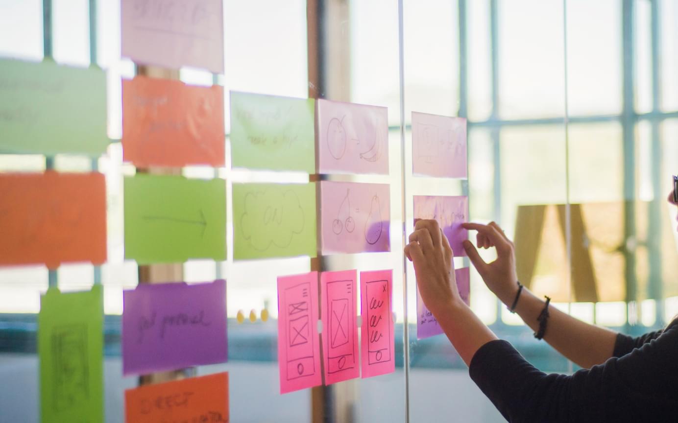 Bunte Post-its werden an einer Glaswand strukturiert angeordnet. Helle, sonnige Atmosphäre.