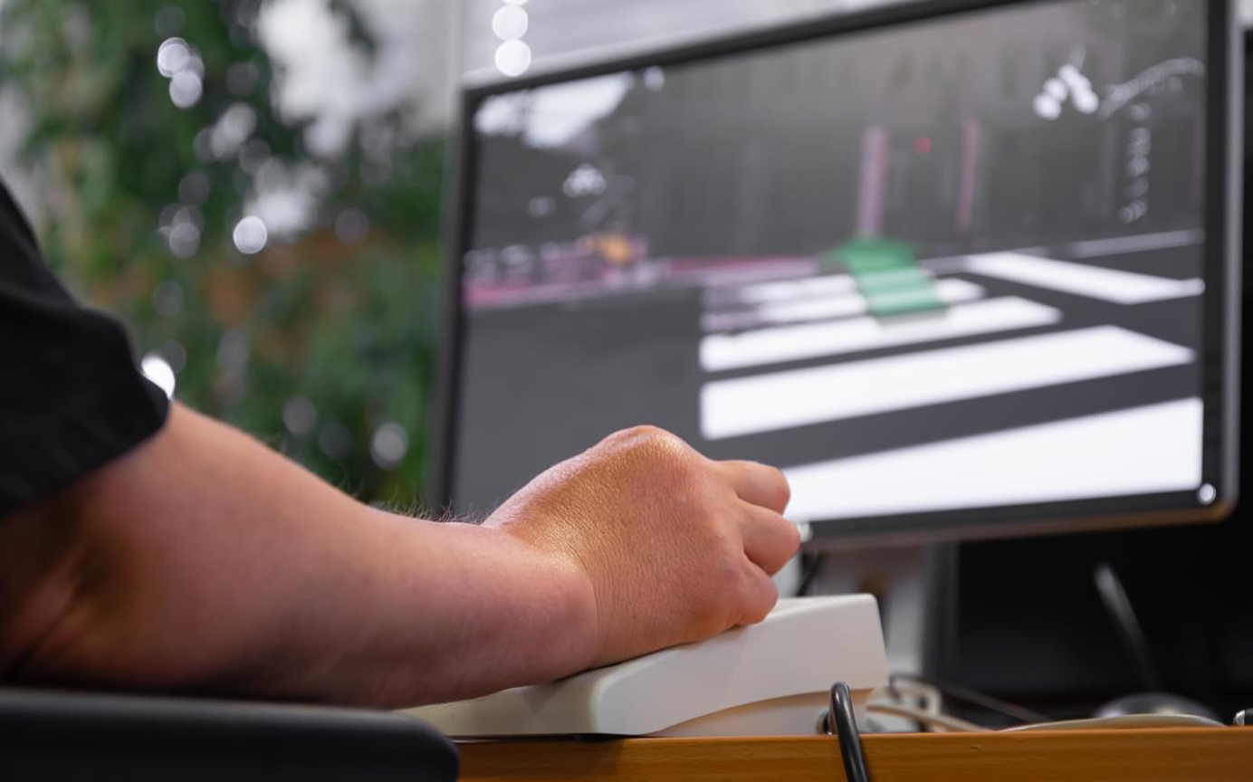 Hand an einem Joystick vor Bildschirm mit Straßen-Simulation – virtuelles Training.

