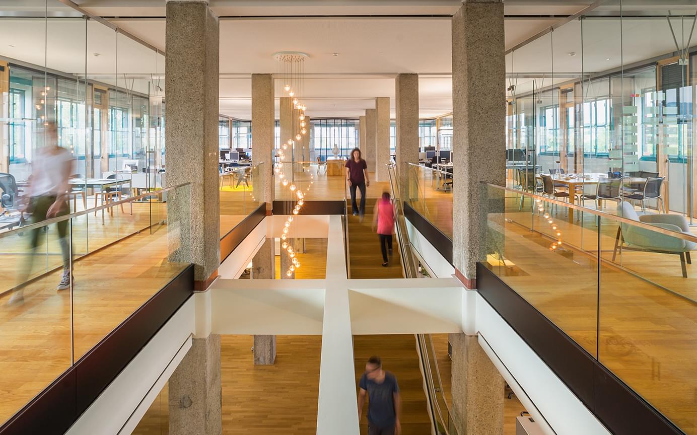 Großzügiges, modernes, sonnendurchflutetes Büro mit zwei Stockwerken, zentraler Treppe Holzfußboden, Glaswänden und stilvoller Hängelampe. Menschen bewegen sich durch das Büro.