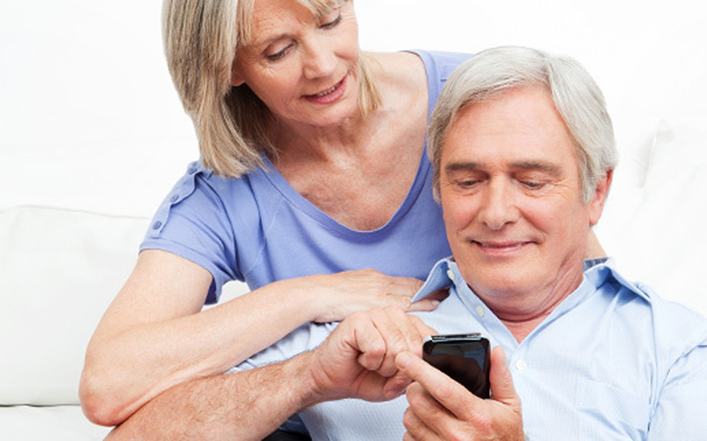 Älteres Paar sitzt auf dem Sofa und bedient gemeinsam ein Smartphone mit einem Lächeln im Gesicht.