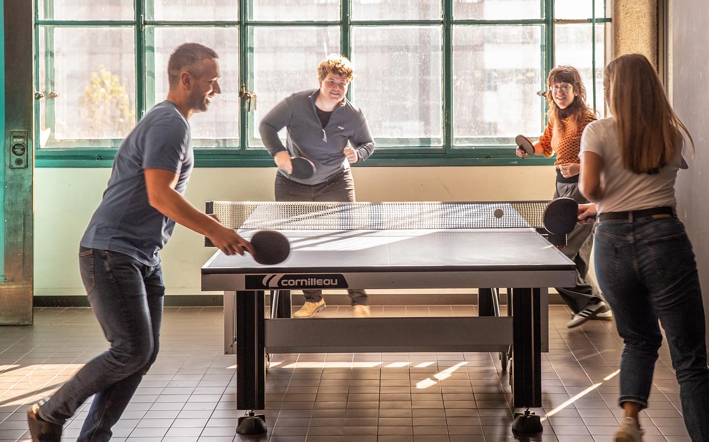 Vier Kolleg:innen spielen Tischtennis in einem sonnigen Freizeitbereich des Büros. Die Stimmung ist fröhlich und dynamisch.