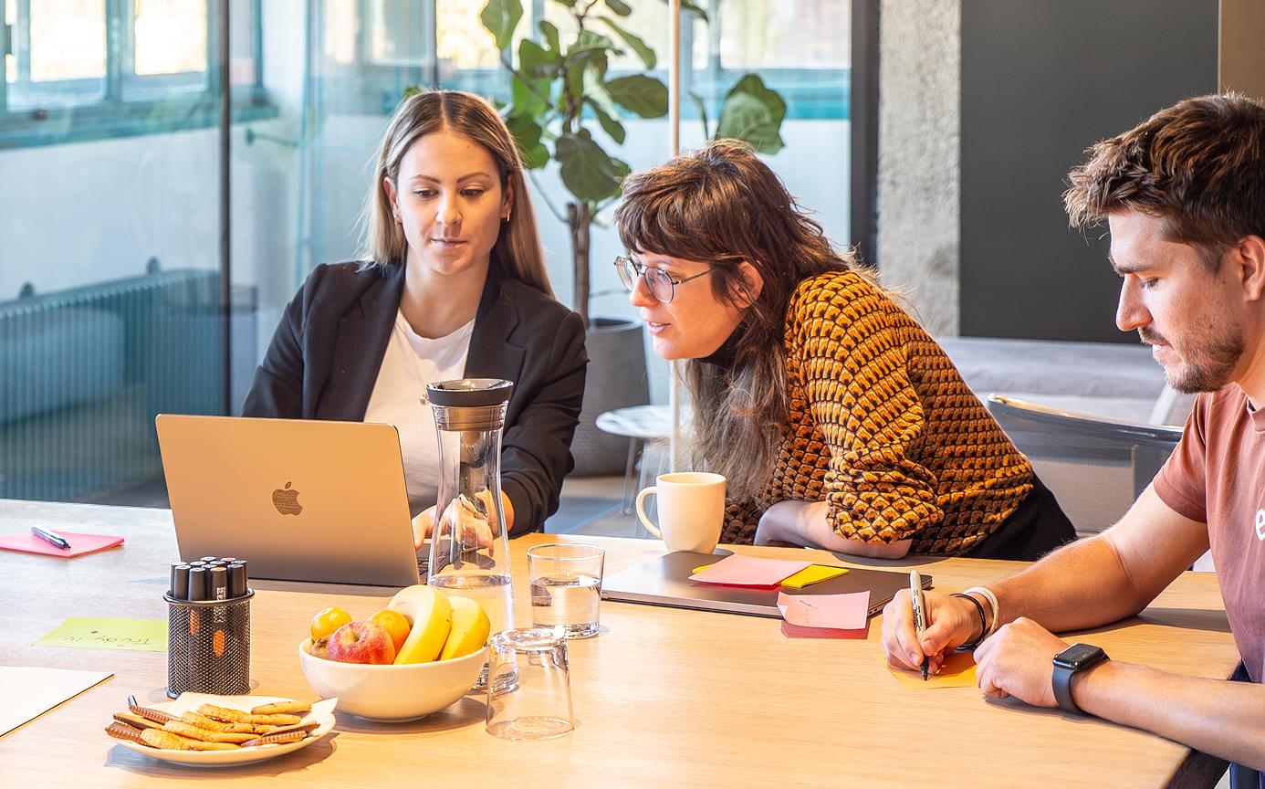 Drei Kolleg:innen sitzen an einem Besprechungstisch und besprechen was sie am Laptopbilschirm sehen. Freundliche, offene Atmosphäre.