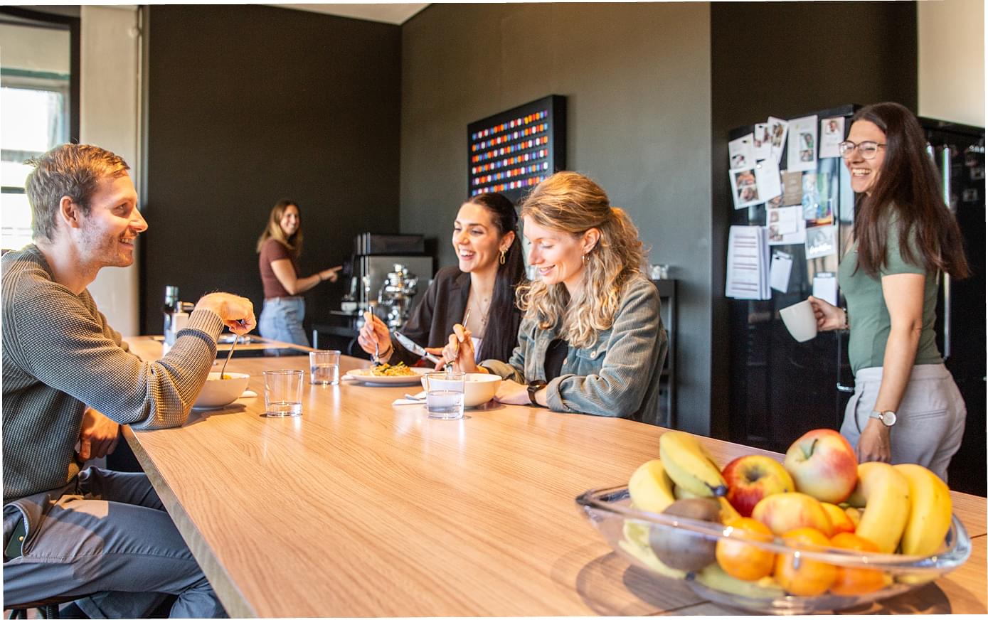 Kolleg:innen sitzen am Mittagstisch mit verschiedenen Speisen und Getränken und einem großen Obstkorb. Eine offene, kommunikative Atmosphäre in der Küche mit weiteren Kolleginnen.