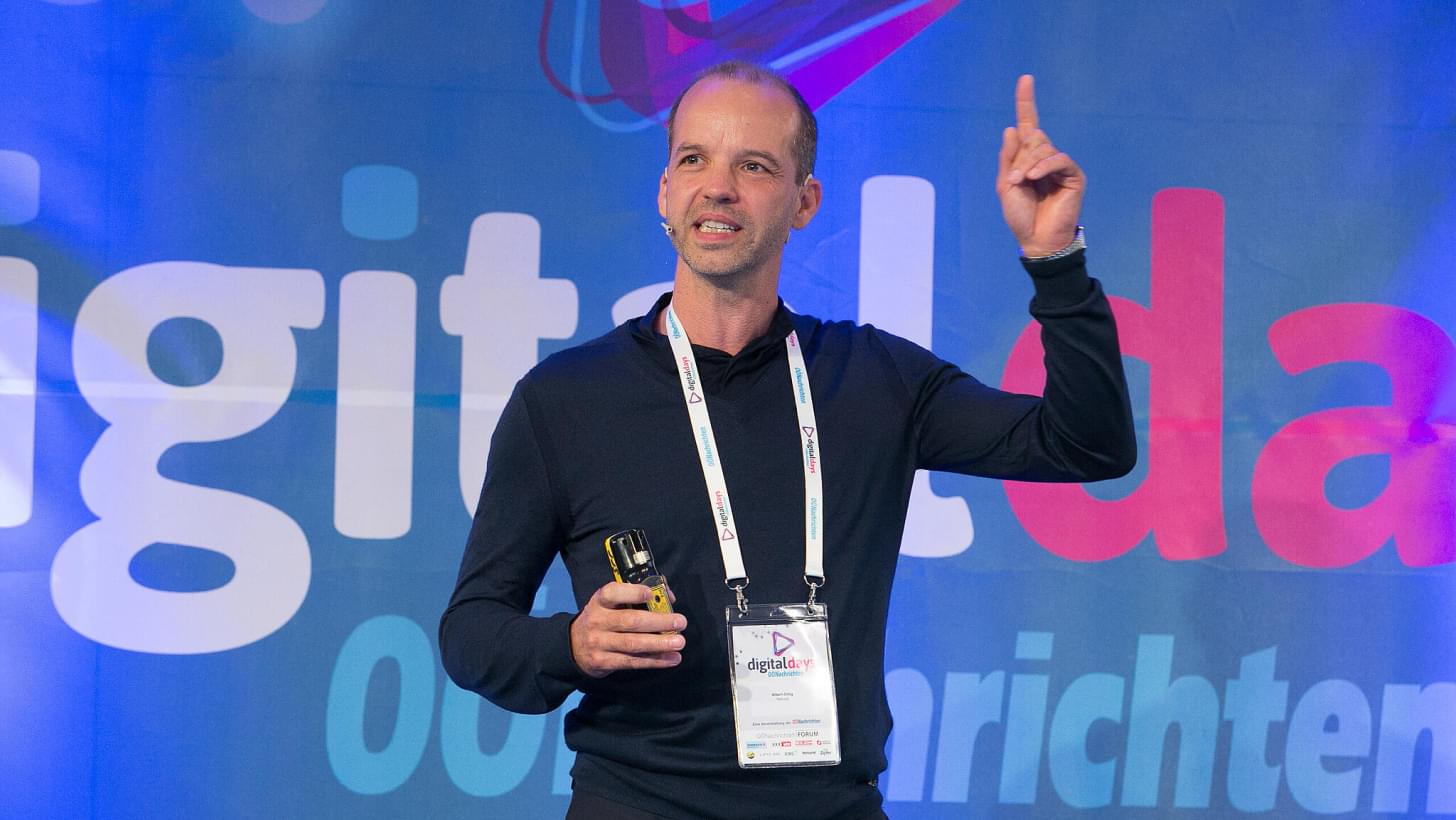 Albert Ortig speaking on stage at Digital Days, holding a presenter and pointing upward.