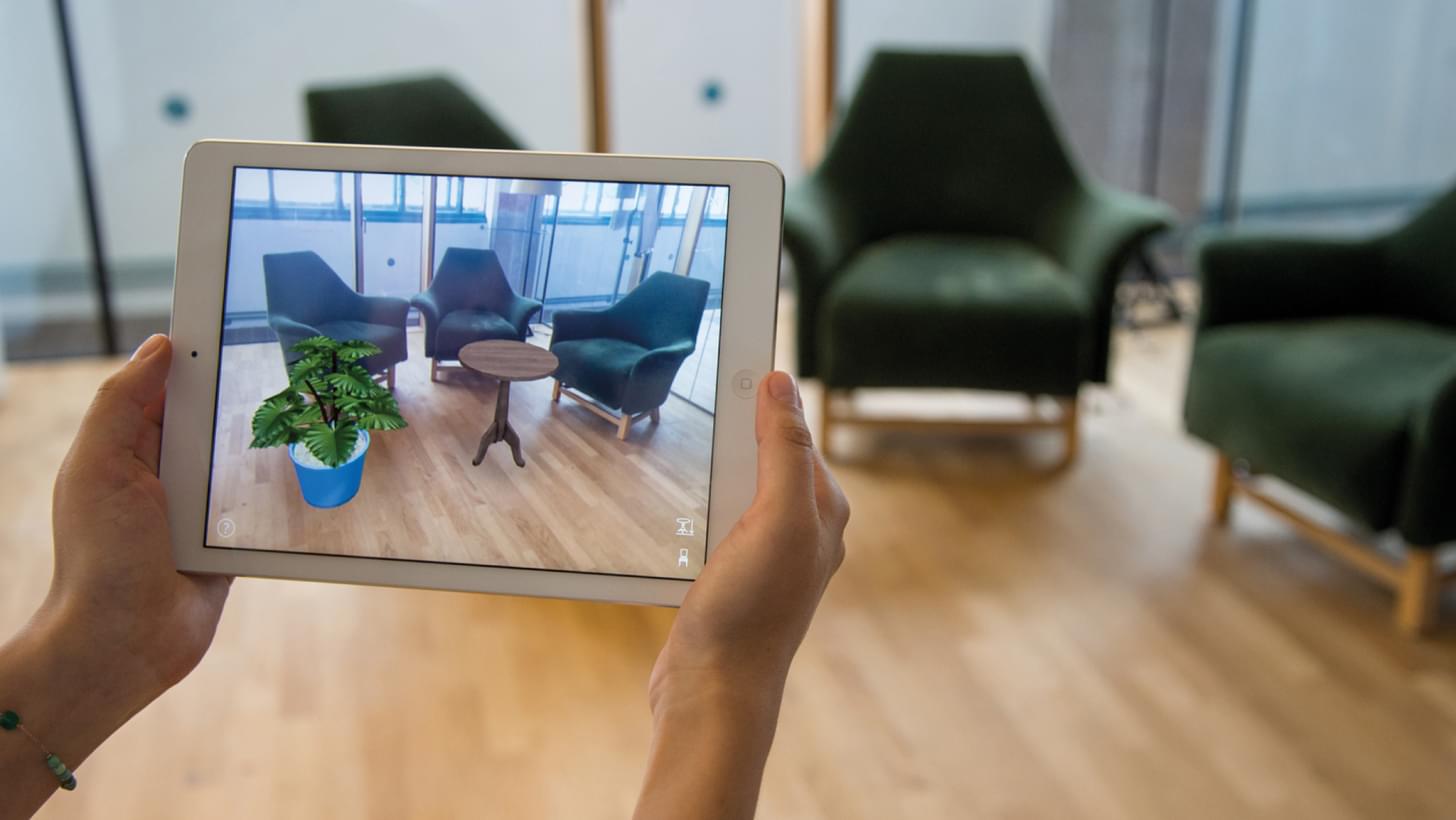 Person hält ein Tablet mit Augmented Reality-Ansicht von Möbeln und Pflanze in einem Raum.