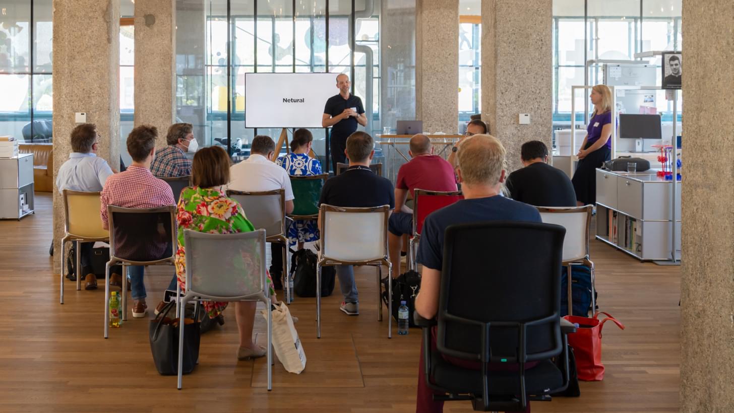 Albert Ortig giving a presentation to a seated audience, screen displays “Netural”.