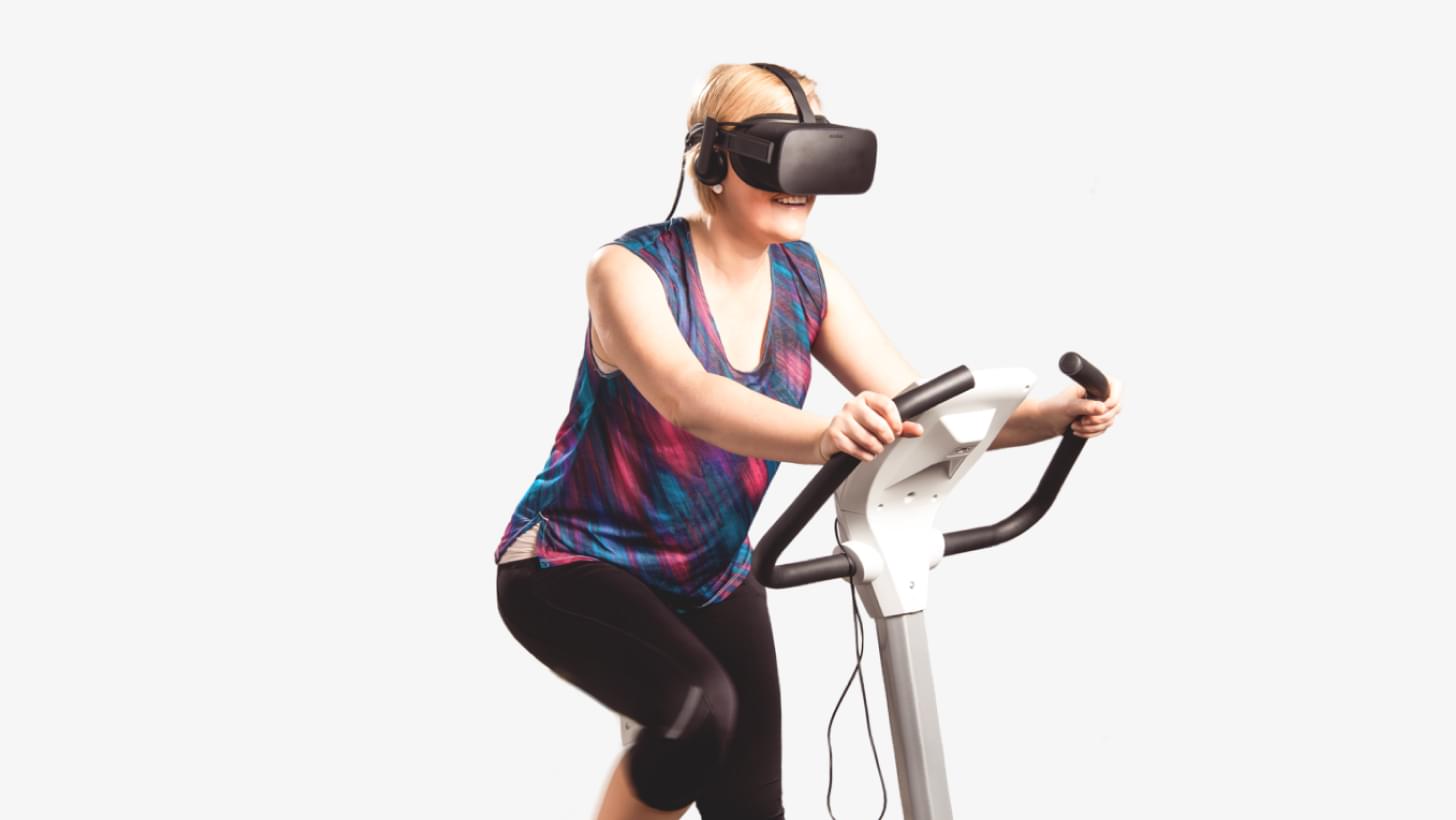 Frau trainiert auf einem Heimtrainer mit VR-Brille und Kopfhörern.
