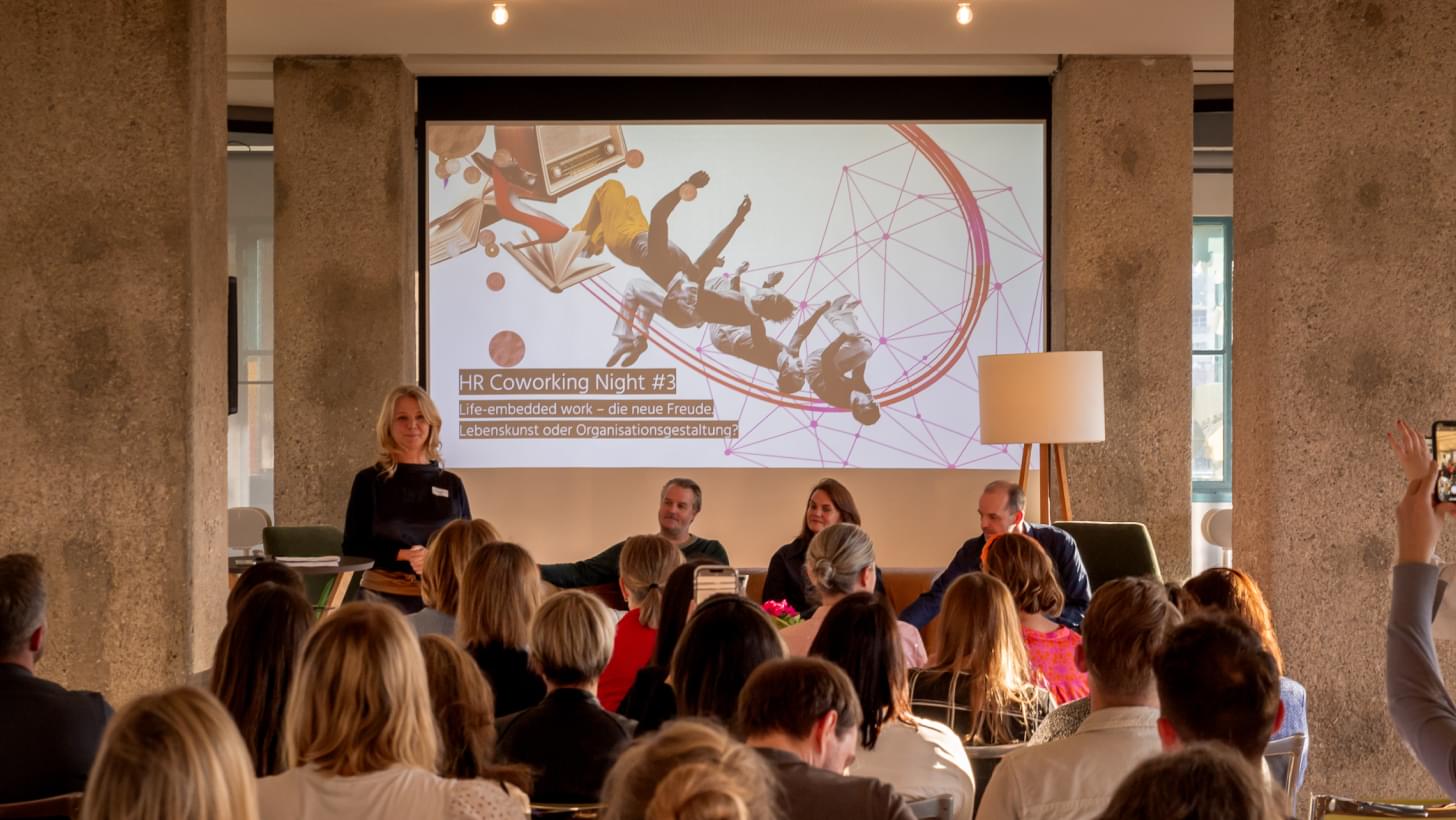 Irene Bouchal-Gahleitner spricht vor Publikum, hinter ihr die Präsentation „Life-embedded work“.