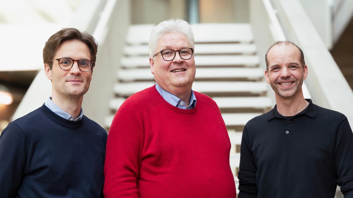 Neusser, Hochreiter und Ortig stehen lächelnd vor einer Treppe in moderner Architektur.