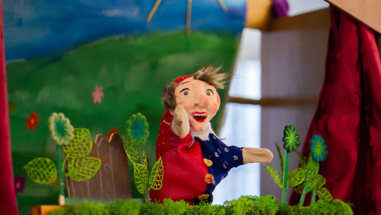 Handpuppe in rotem Kostüm auf bunt dekorierter Puppenbühne.