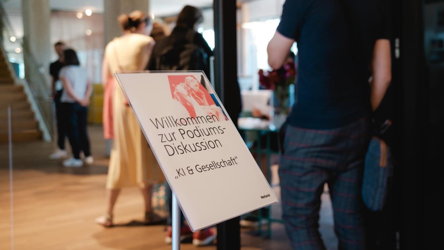 Aufsteller mit der Aufschrift „Willkommen zur Podiumsdiskussion – KI & Gesellschaft“, Besucher betreten die Location.