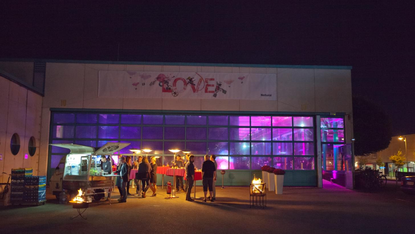 People in front of the illuminated Tabakfabrik Linz at night; banner with "LOVE" over the entrance.