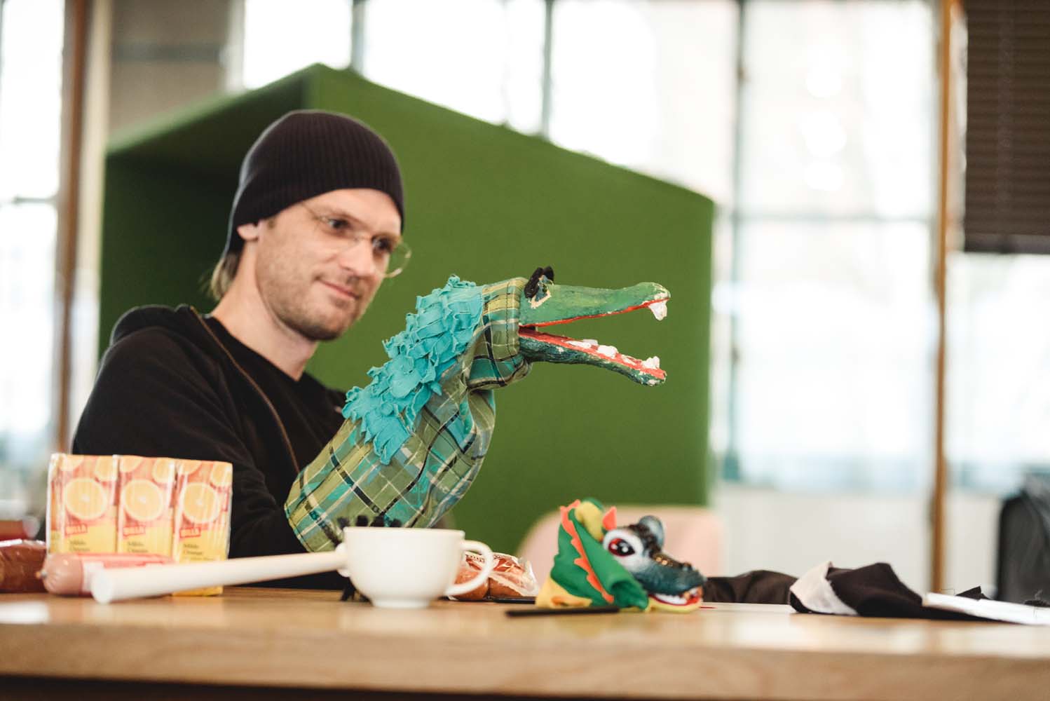 Mann sitzt am Tisch und spielt mit einer grünen Krokodil-Handpuppe.