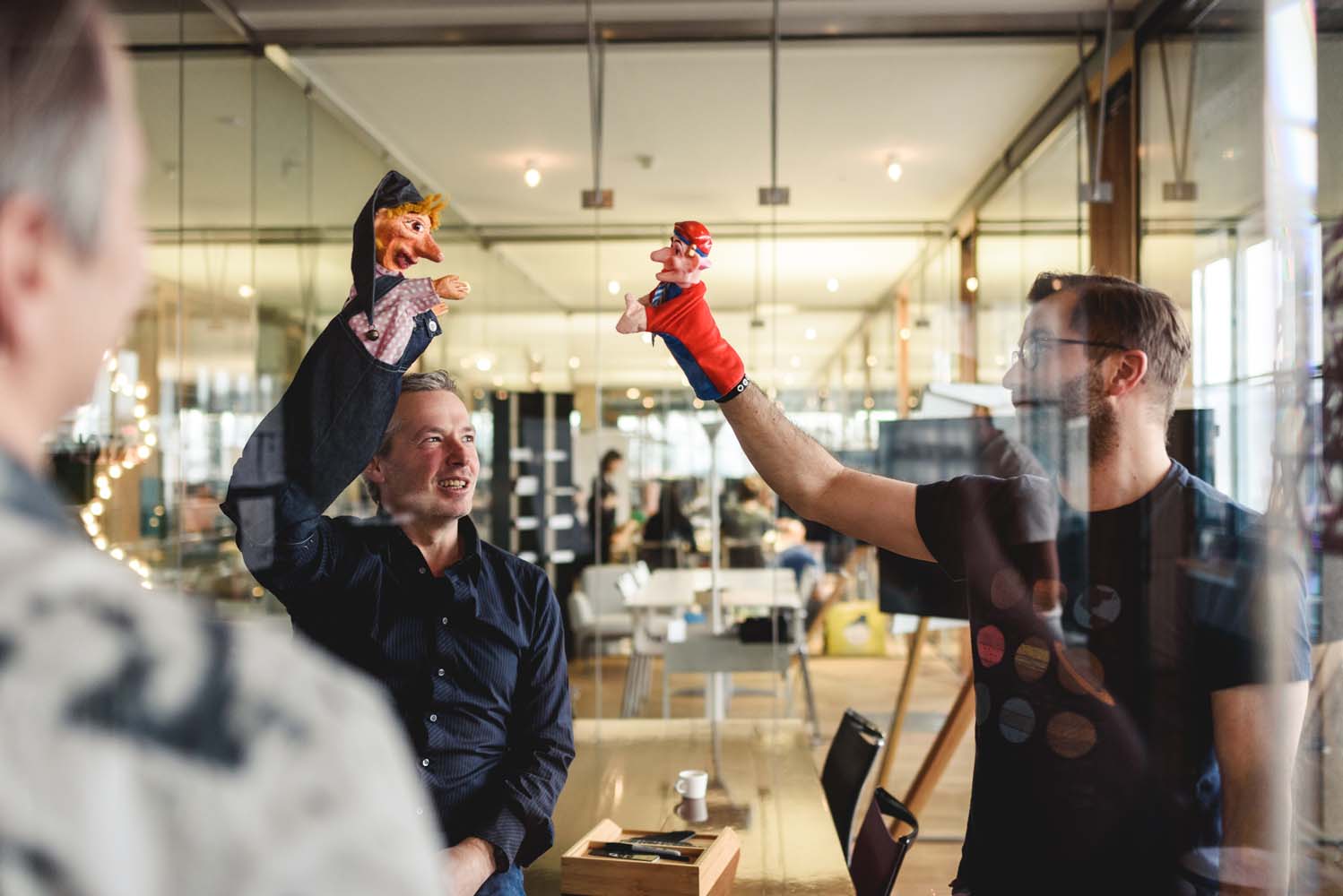 Zwei Männer spielen mit Kasperle-Fingerpuppen in einem modernen Büroraum.