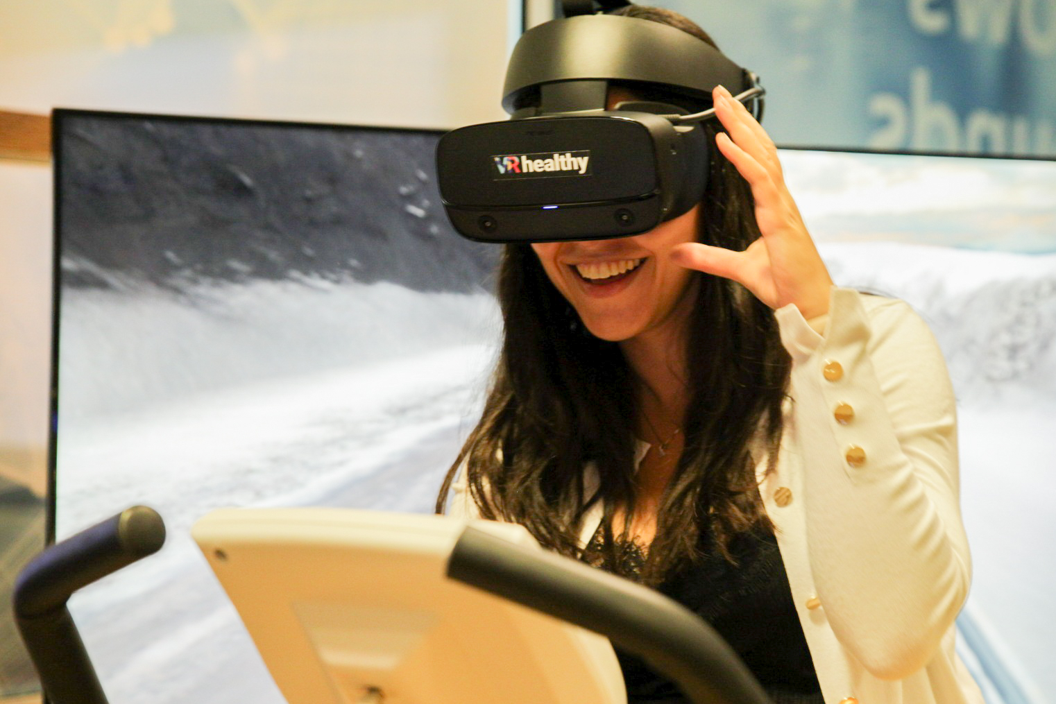 Frau nutzt Heimtrainer mit VR-Brille „VRhealthy“ vor Bildschirm mit Landschaftsszene.
