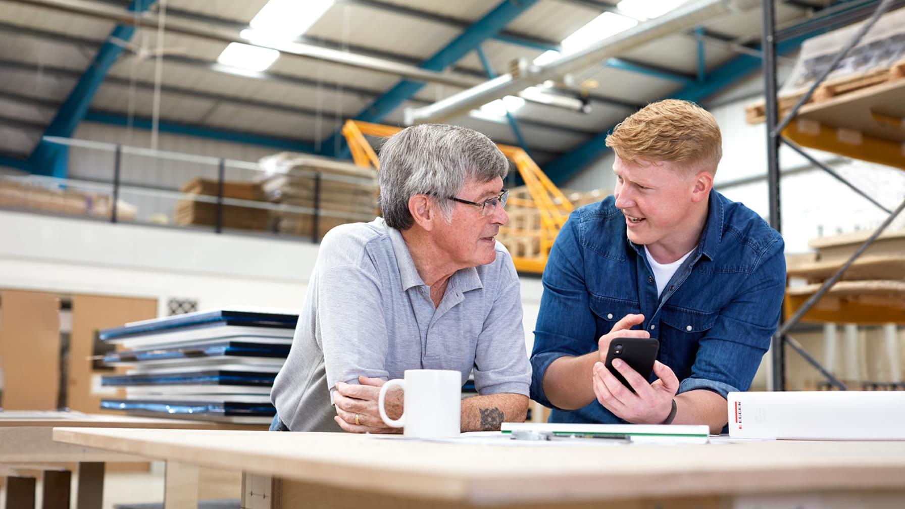 Zwei Männer lächeln und unterhalten sich in einer Werkstattumgebung über etwas auf einem Smartphone; im Hintergrund sind Werkzeuge und Materialien zu sehen.