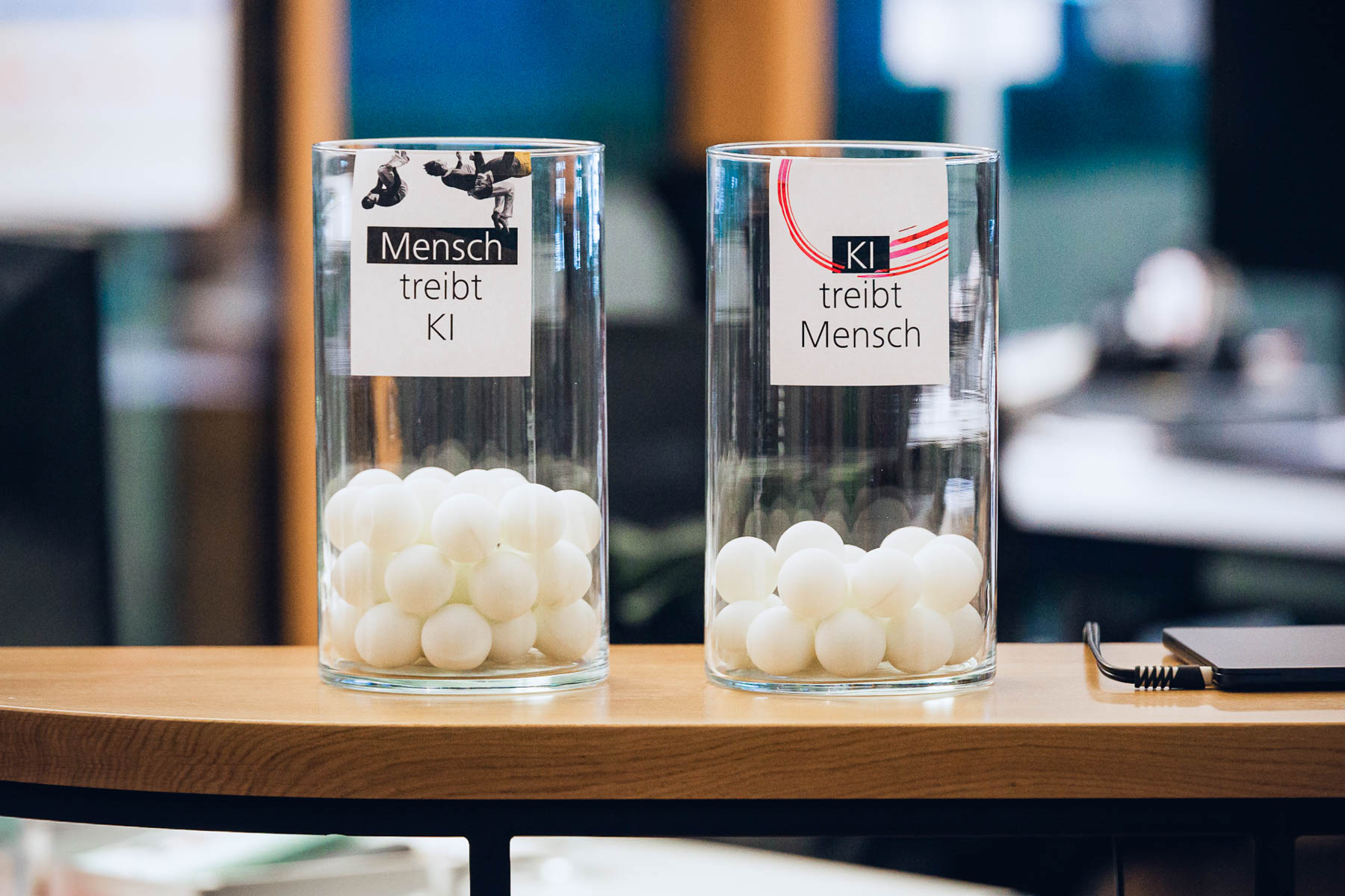 Zwei Glasgefäße mit weißen Kugeln, beschriftet mit „Mensch treibt KI“ und „KI treibt Mensch“, Publikumsabstimmung.
