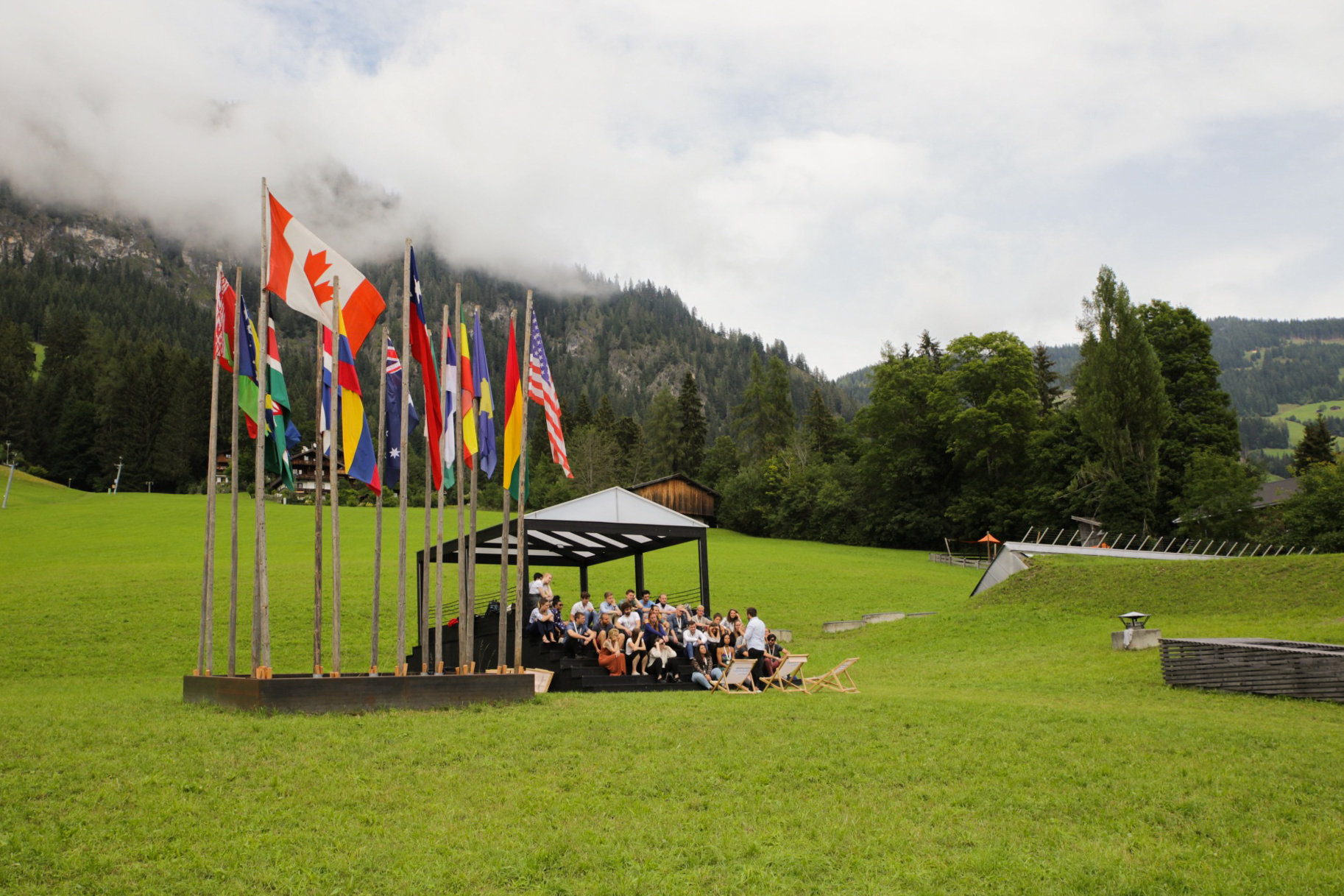 Open-Air-Veranstaltung mit internationalen Flaggen auf grüner Wiese vor Bergkulisse.