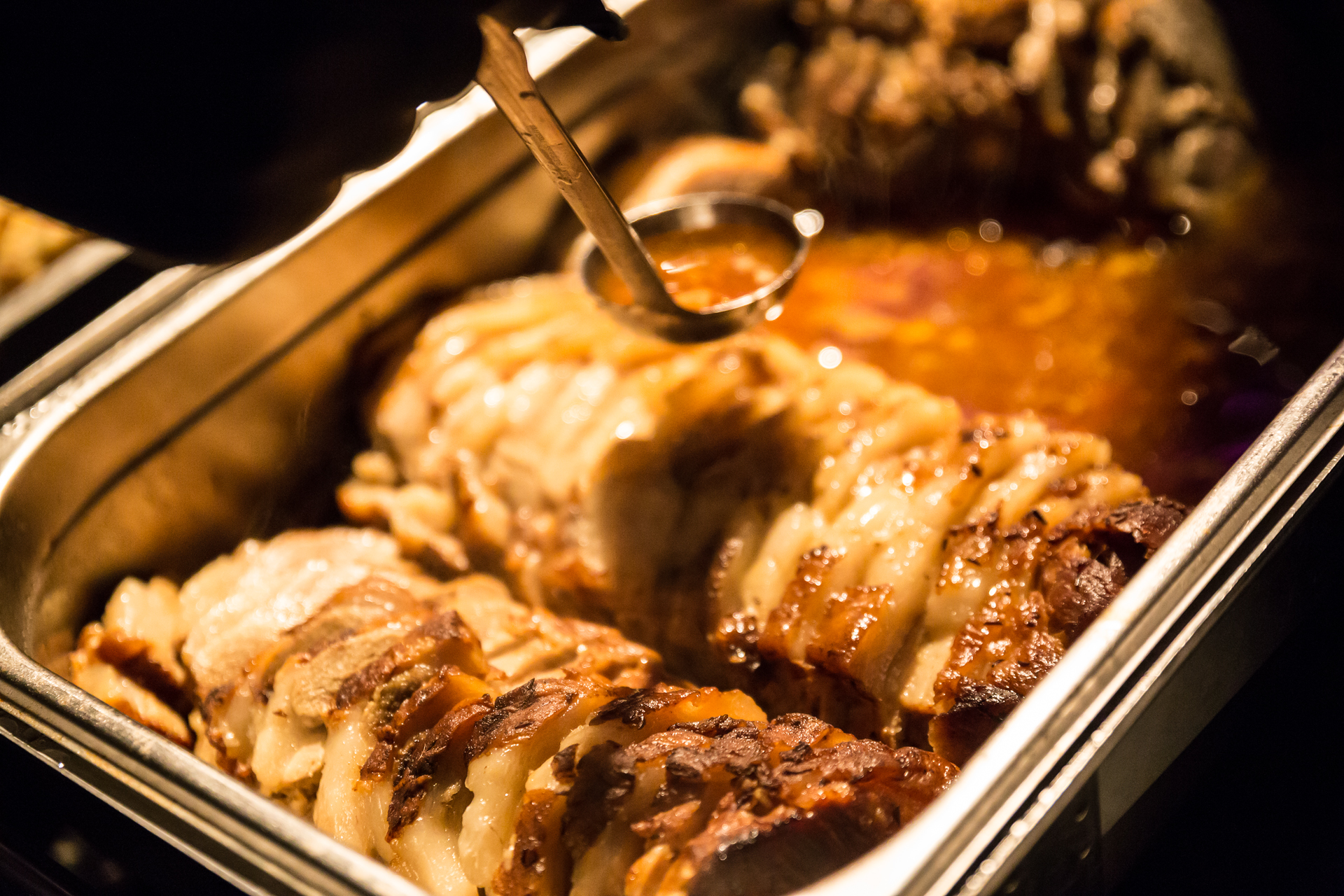 Roasted meat pieces in a warming container are being covered with sauce.