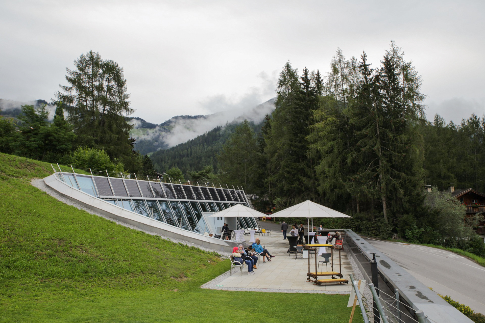 Terrasse mit Sonnenschirmen und Solaranlage, umgeben von Wald und Bergen.