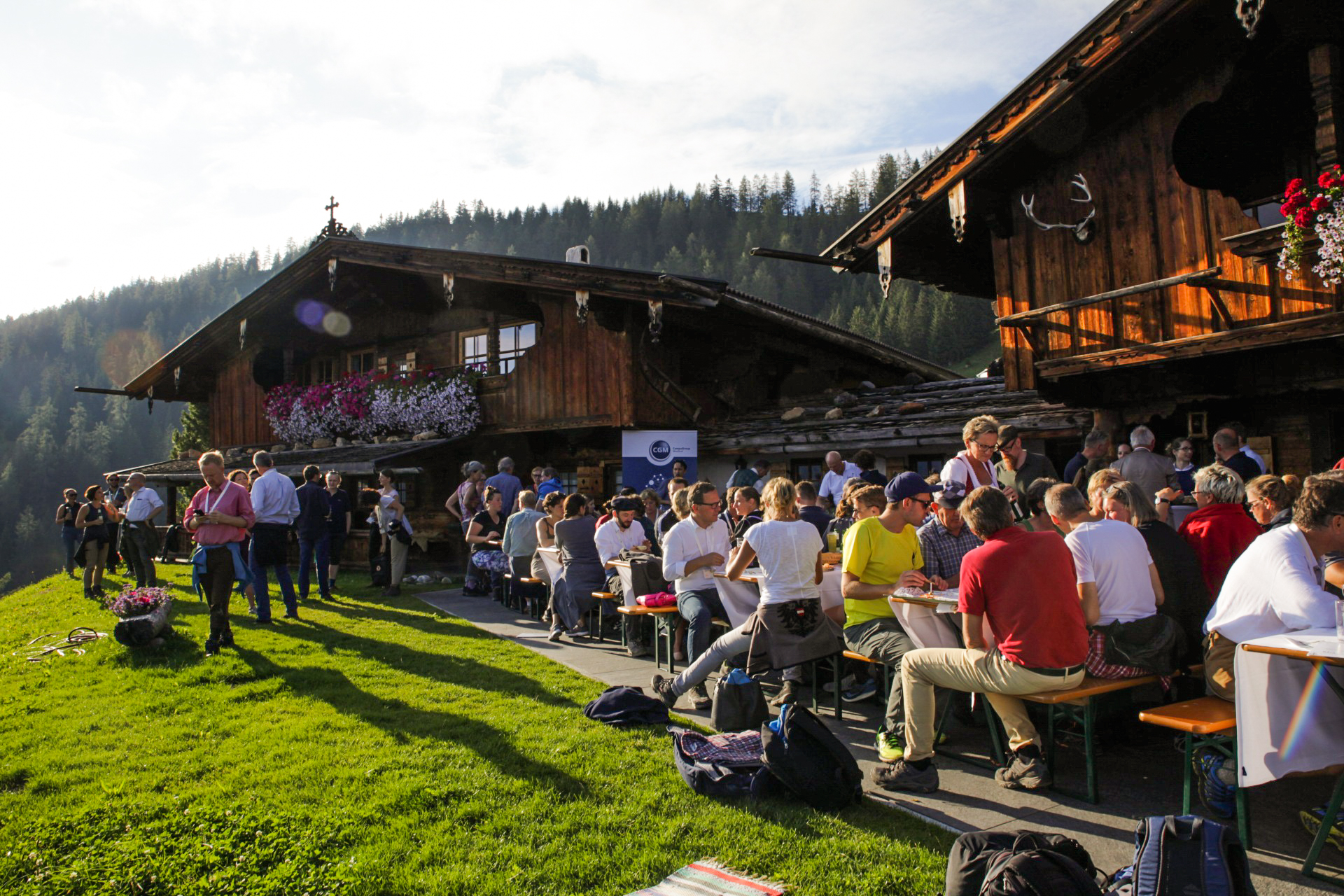 Menschen sitzen an Tischen vor einer Almhütte bei sonnigem Wetter.