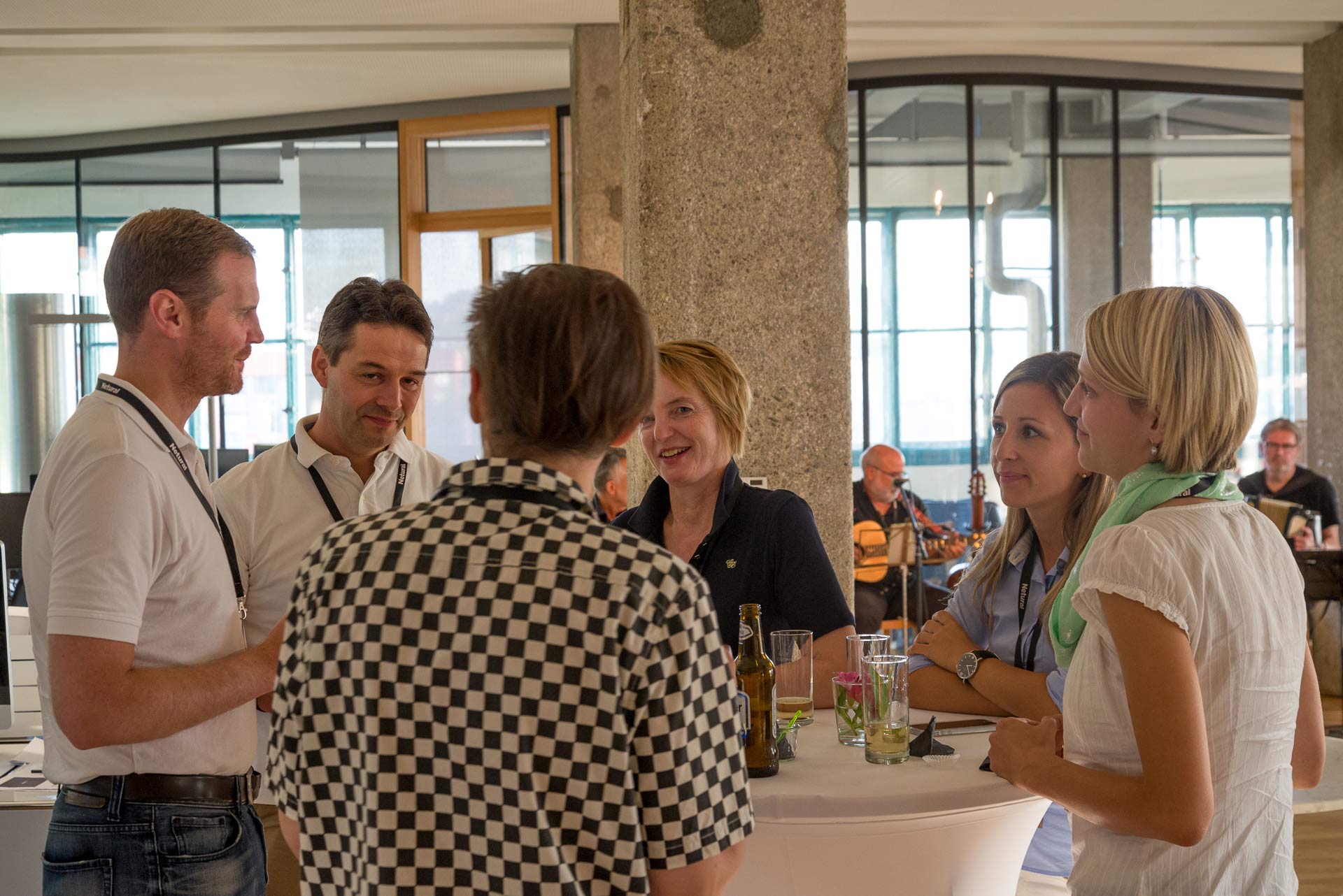 Several people are standing around a round table with drinks, engaging in conversation.
