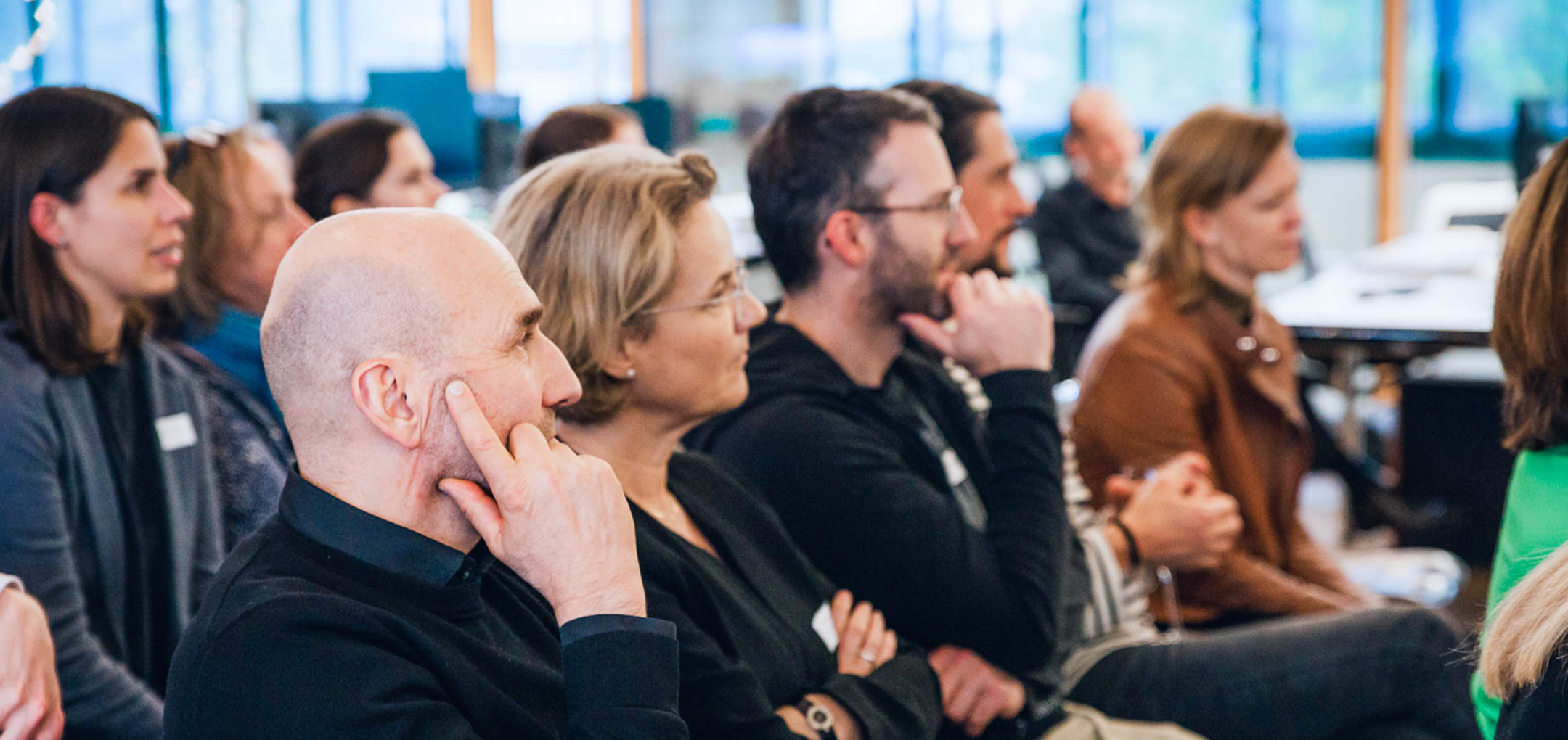 Teilnehmer:innen sitzen in einer Reihe und hören aufmerksam einer Präsentation zu– Fokus und Nachdenklichkeit prägen den Moment.