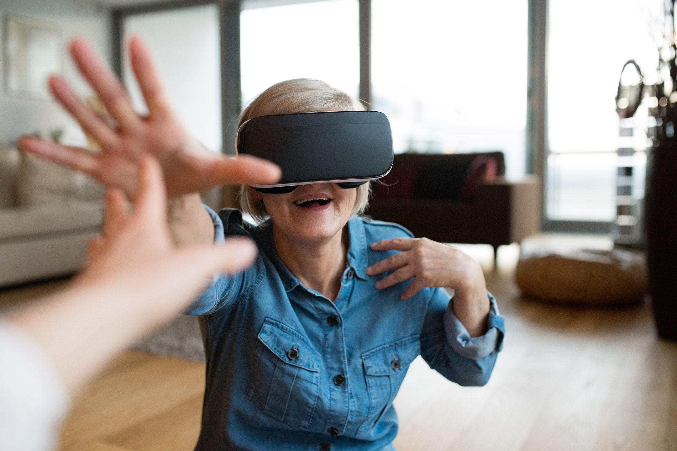 Eine ältere Dame trägt eine VR-Brille, sie lacht und berührt die Hand einer Person hinter der Kamera.