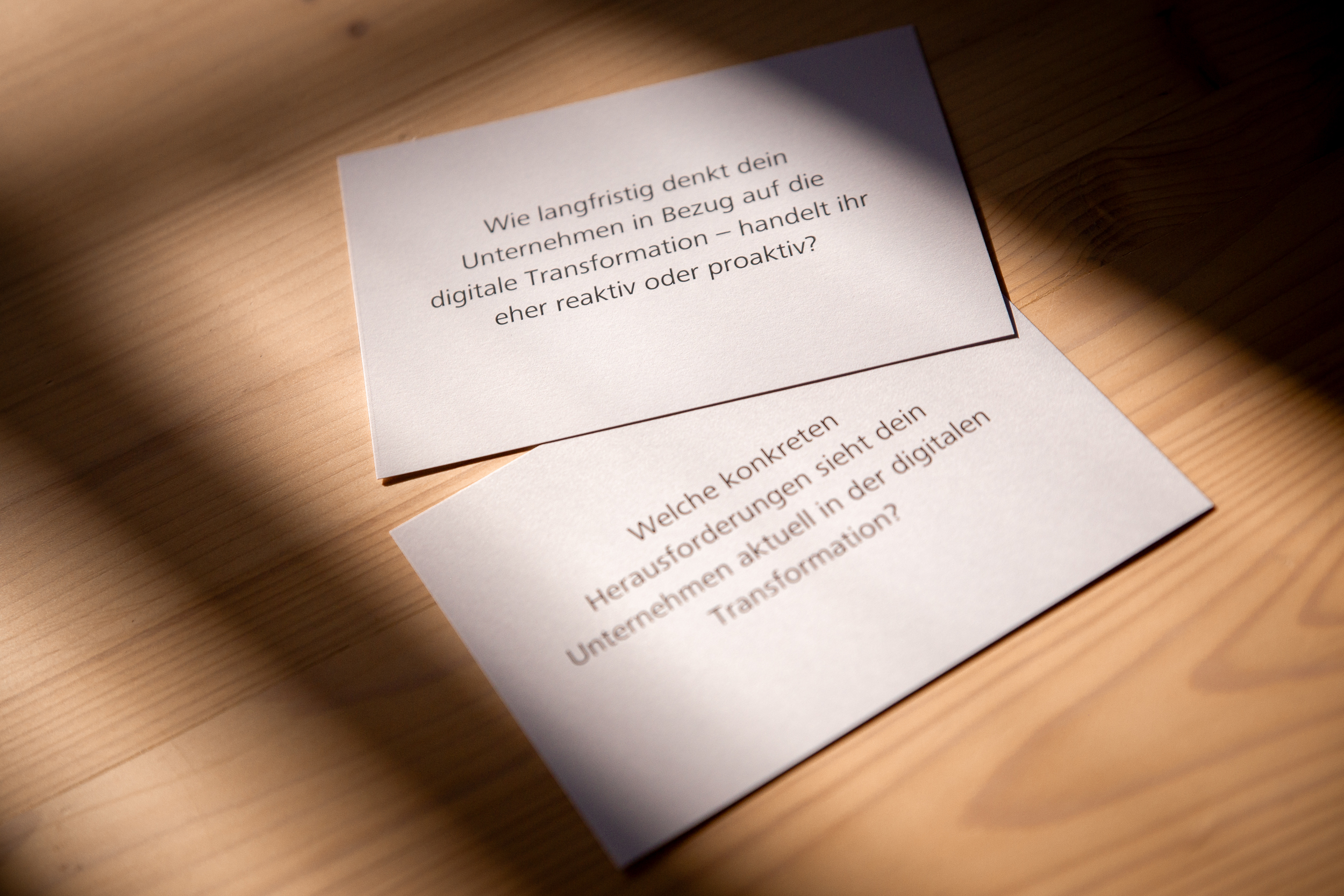 Two cards with reflection questions on digital transformation placed on a wooden table, lit by angled sunlight.