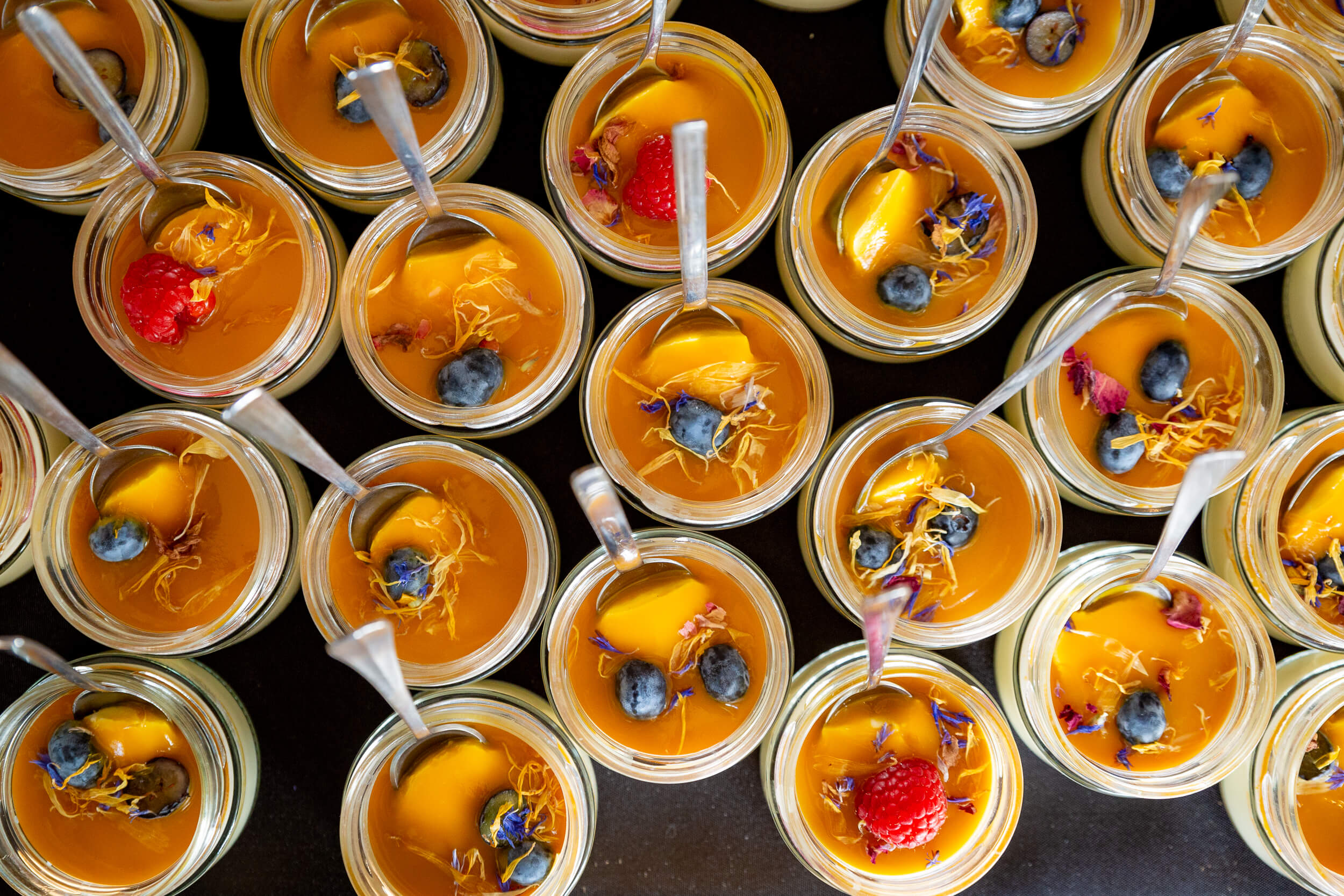 Viele Dessertgläser mit gelber Creme, Beeren und Blüten, samt Löffeln auf schwarzem Hintergrund arrangiert.