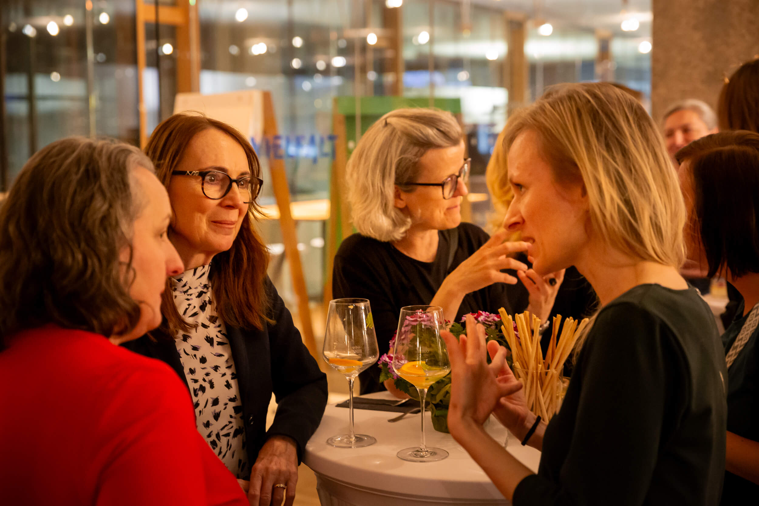 Frauen in lebhaftem Gespräch an einem kleinen Stehtisch mit Getränken und Blumendekoration in einem stimmungsvoll beleuchteten Raum.