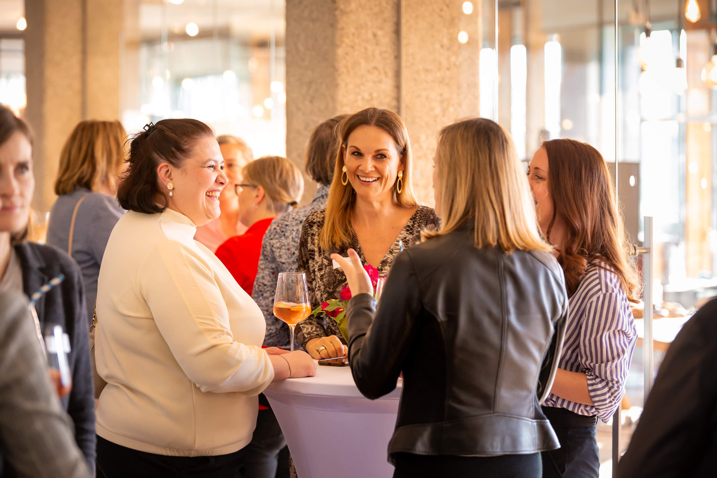 Frauen stehen an einem Stehtisch, lachen und unterhalten sich entspannt bei einem Drink.