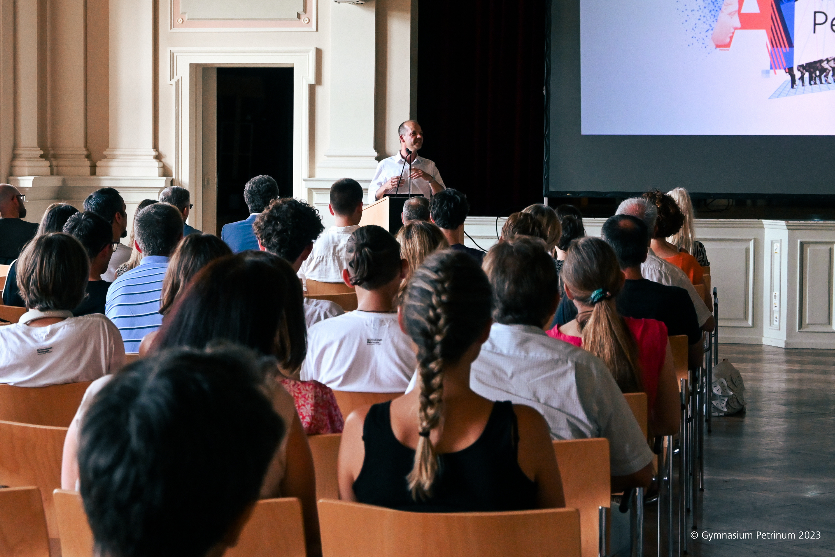 Albert Ortig spricht in der Aula des Petrinum Linz, im Vordergrund sitzen Eltern oder Gäste auf Holzstühlen.