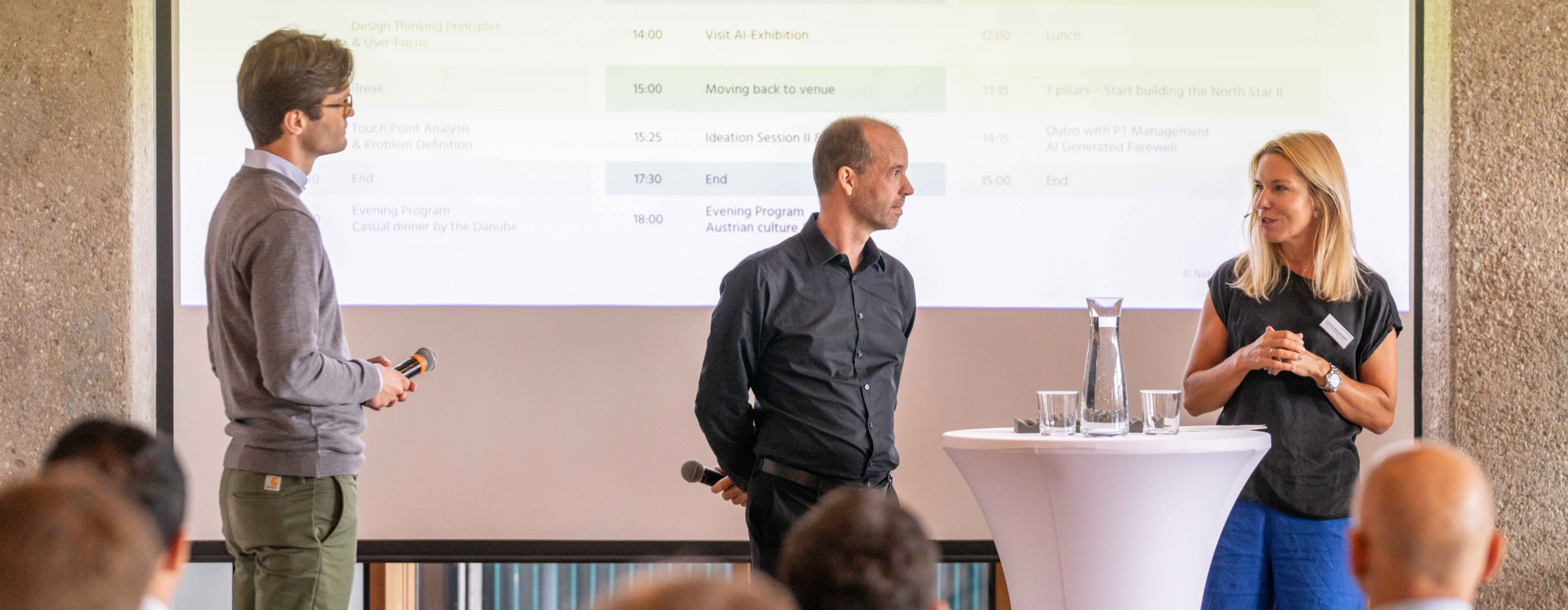 Drei Vortragende diskutieren auf einer Bühne mit Tagesagenda im Hintergrund.