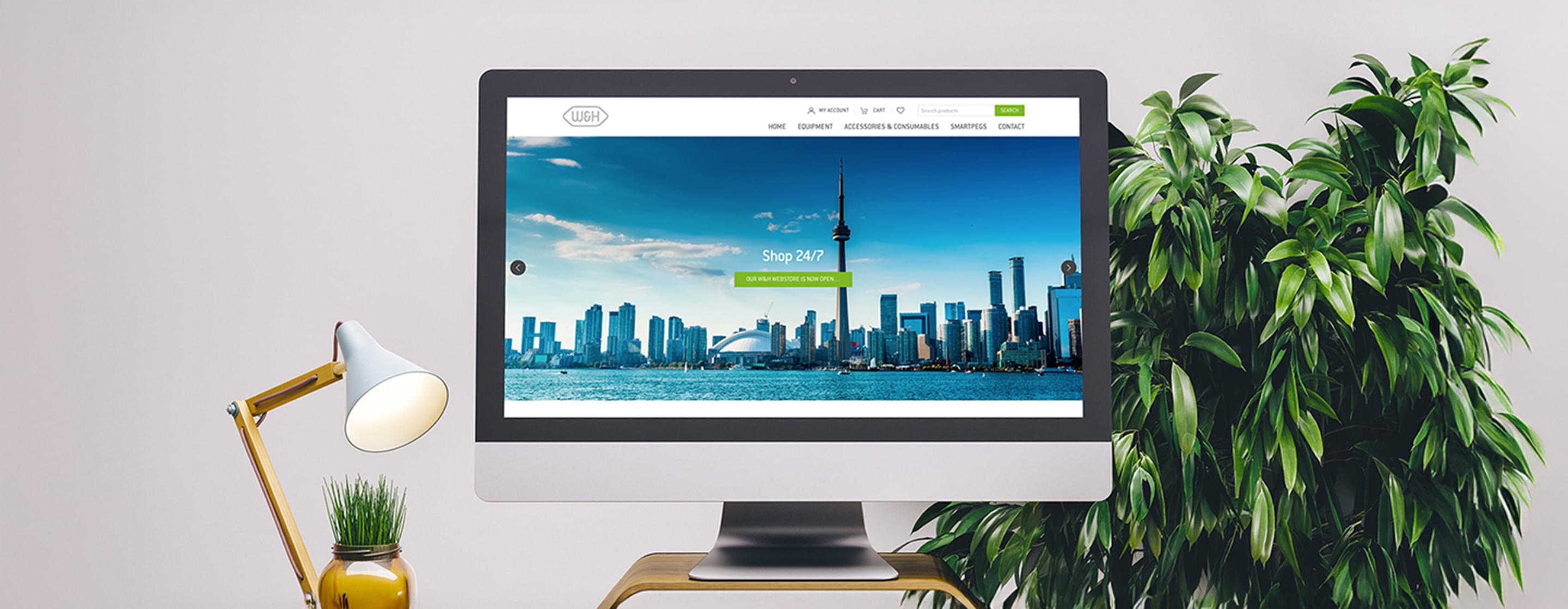 Monitor mit geöffneter W&H-Webseite, davor Lampe und Pflanze auf einem hellen Schreibtisch.