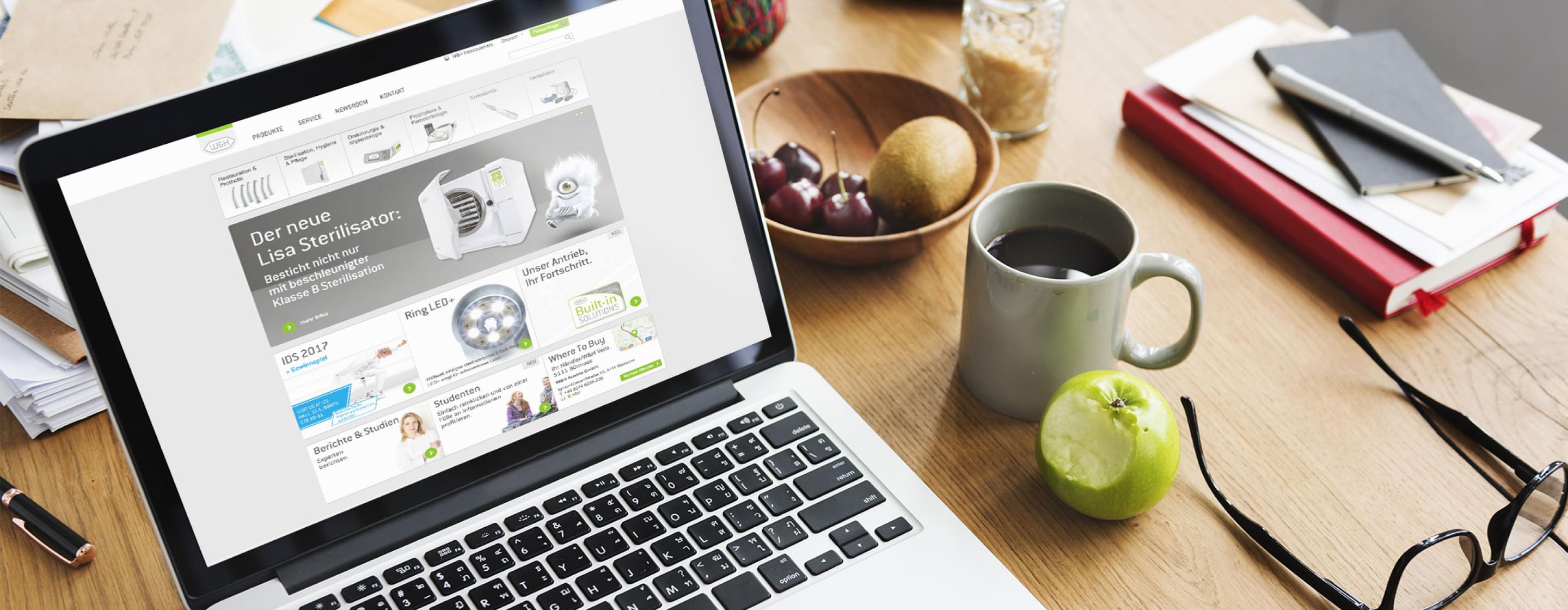 Laptop mit geöffneter W&H-Webseite, daneben Kaffee, Apfel, Brille und Notizbücher auf Holztisch.