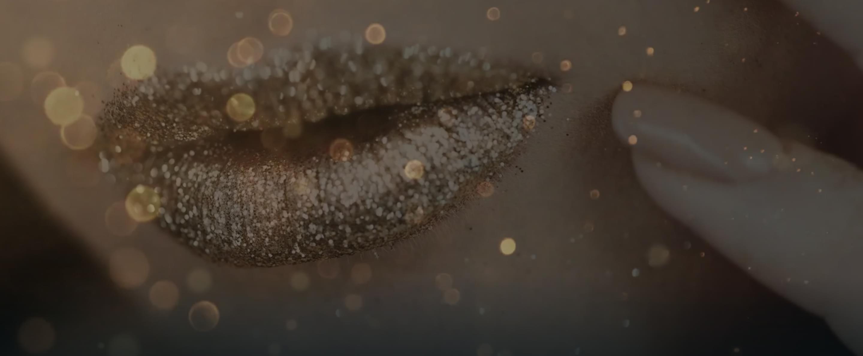 Nahaufnahme von Lippen mit goldenem Glitzer Make-up, umgeben von funkelnden Lichtpunkten.