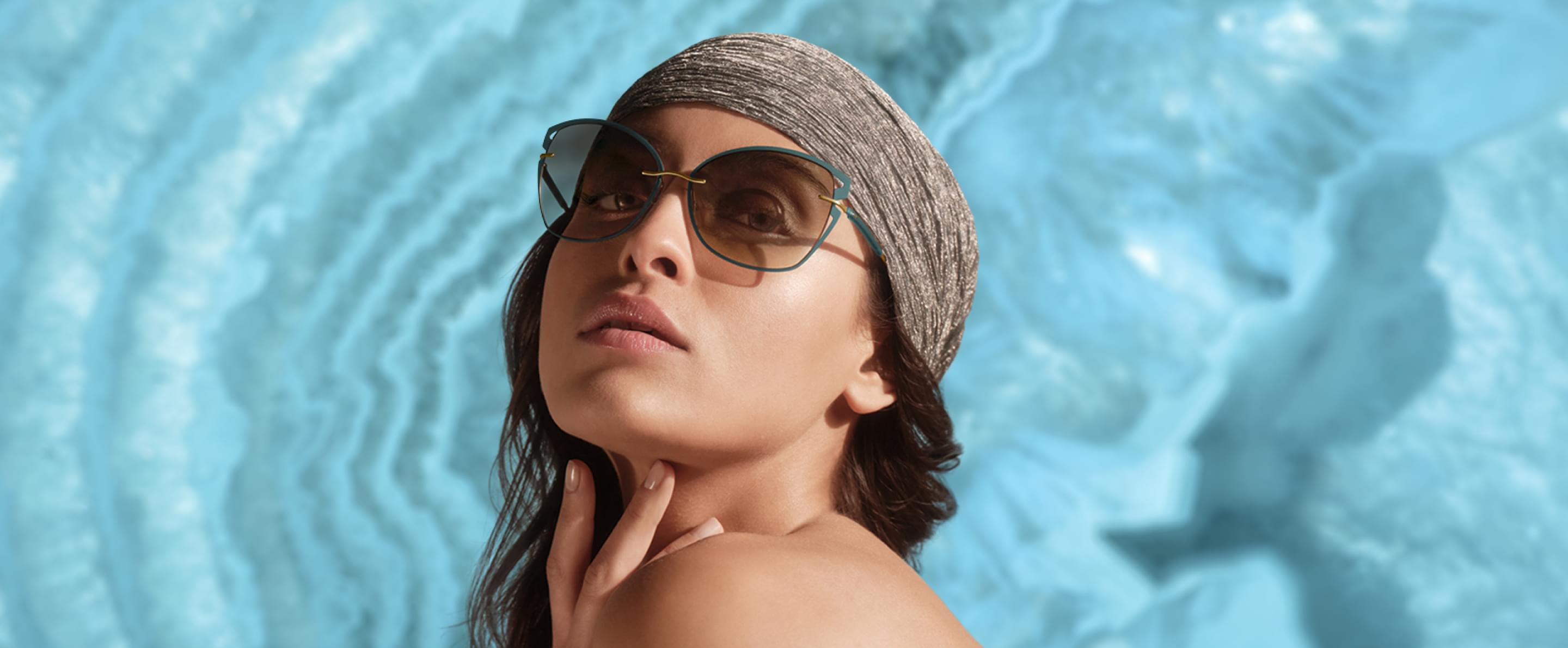 Eine Frau mit Kopftuch und stilvoller Silhouette Sonnenbrille blickt direkt in die Kamera, vor blauem Hintergrund.