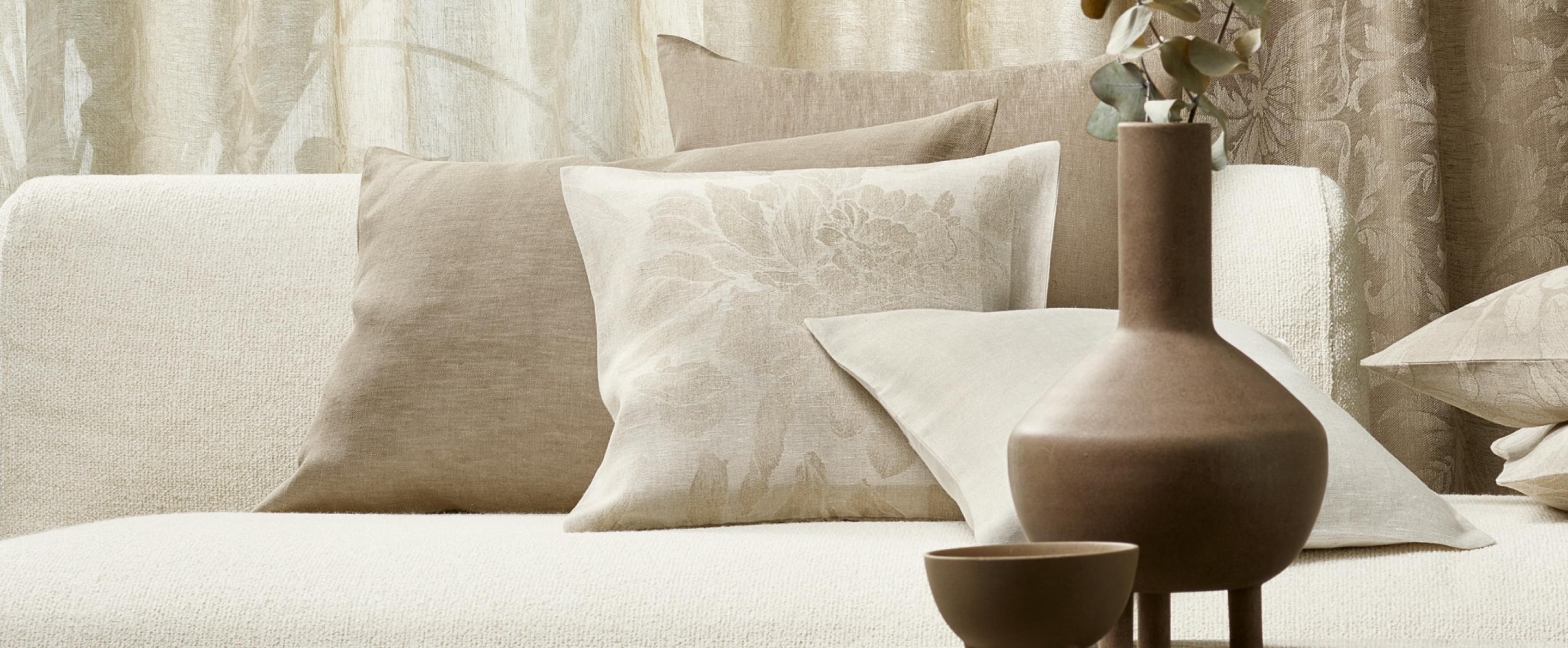 Beige Sofakissen mit floralen Mustern und Leinenstruktur, ergänzt durch braune Keramikvase im Vordergrund.