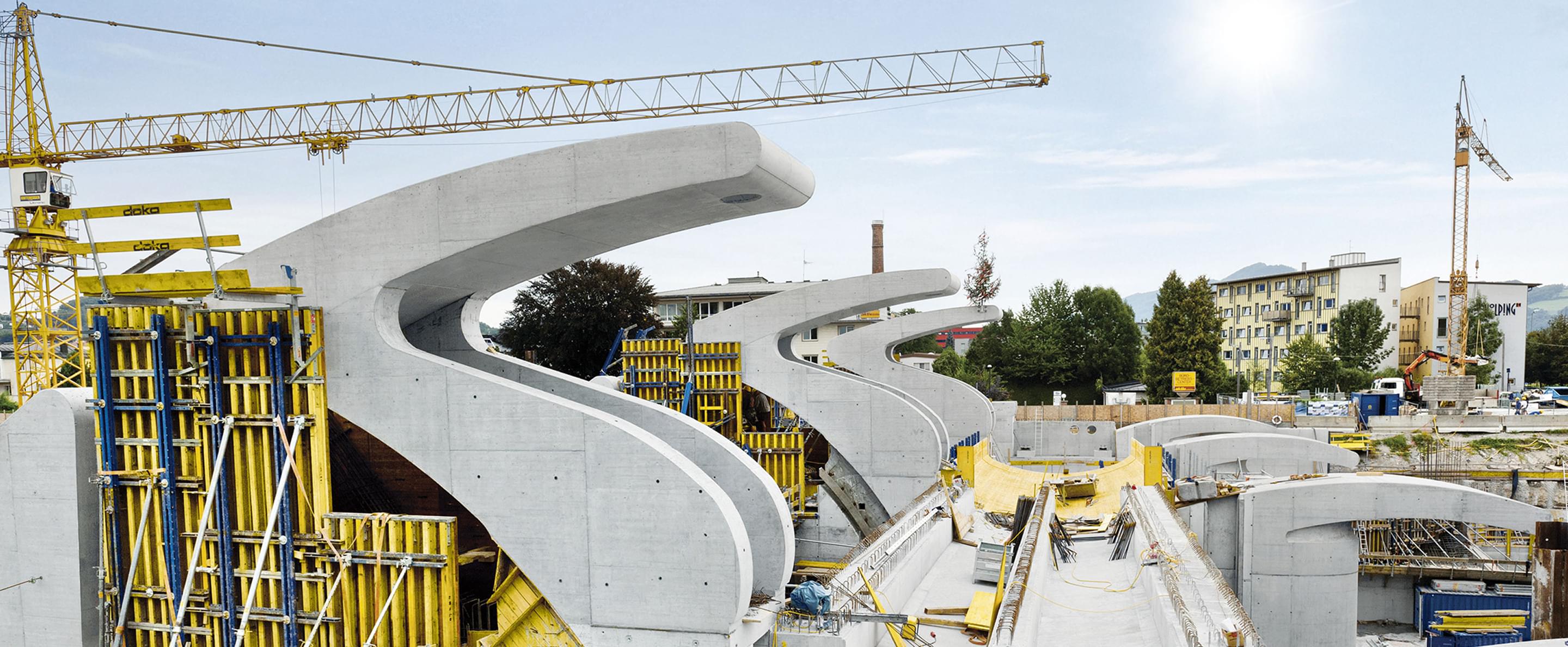 Brückenbau mit geschwungenen Betonelementen und gelbem Kran bei Sonnenschein, im Hintergrund Wohn- und Bürogebäude.