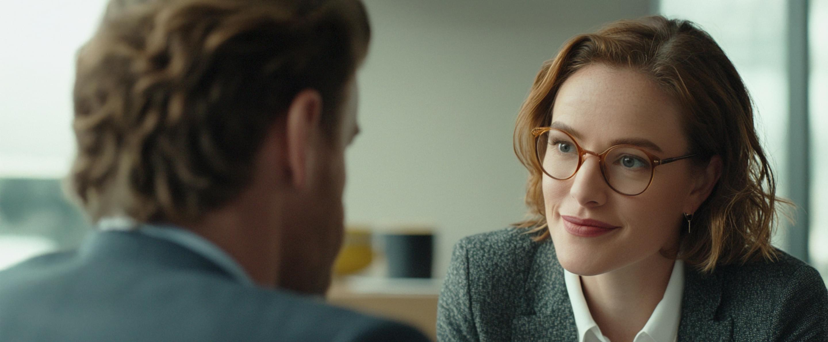 Frau mit Brille lächelt einem Mann im Anzug zu, beide in einem Gespräch in professioneller Umgebung.