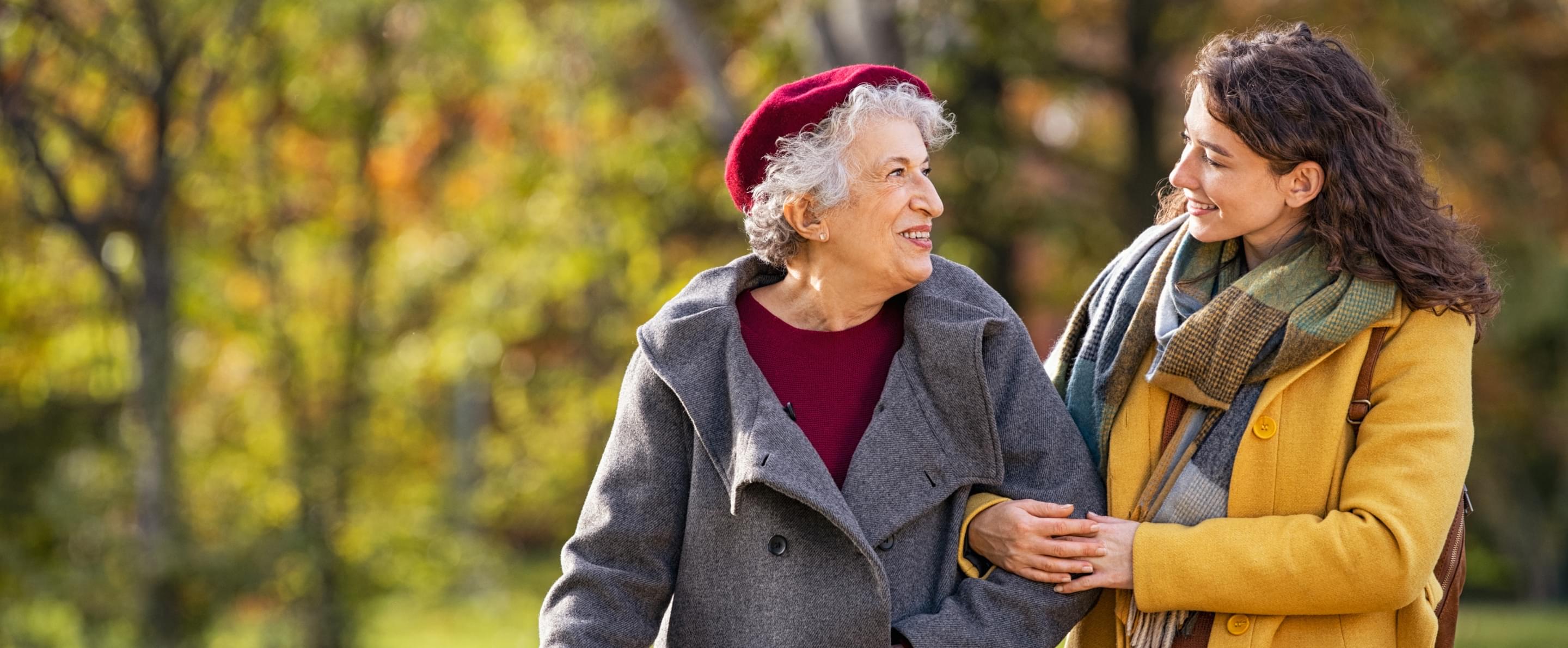 Junge Frau spaziert lachend mit älterer Frau durch herbstlichen Park, beide warm gekleidet und arm in arm.