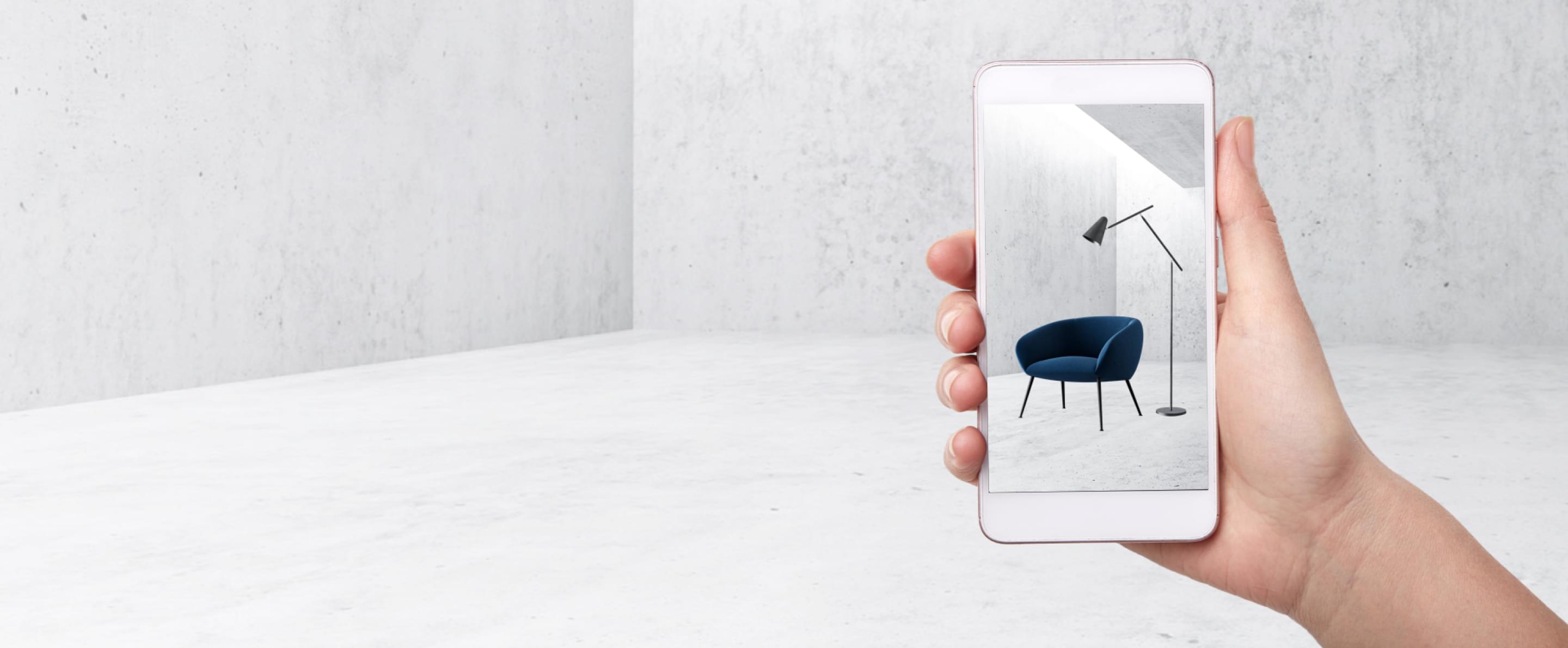 Hand hält Smartphone mit AR-Ansicht eines blauen Sessels und einer Stehlampe in minimalistischem Raum.